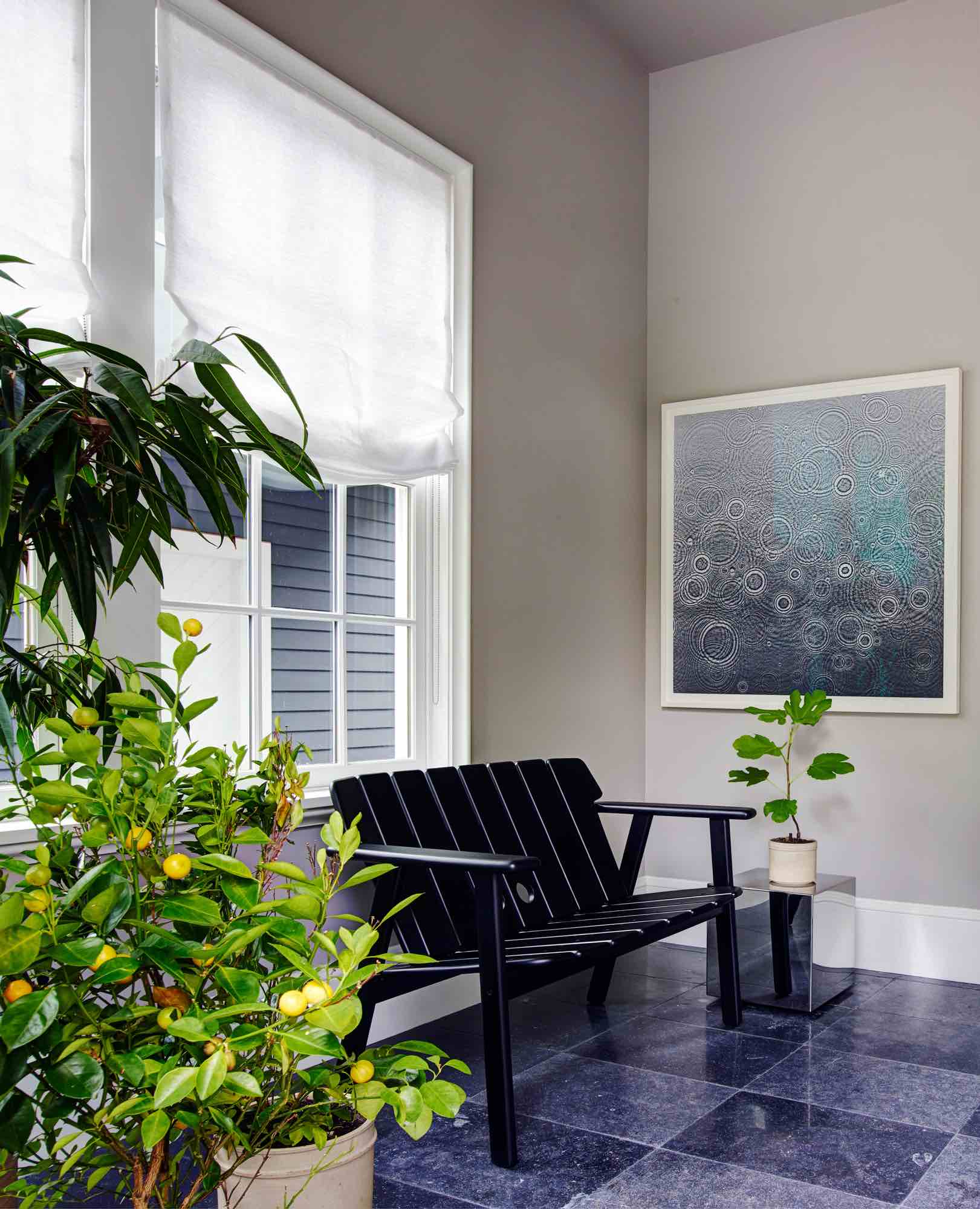 In the mudroom designed by Carol Egan, Belgian block slab and honed floor is contrasted by grey painted walls.  An ebonized slatted bench by Sergio Rodrigues sits under the window flanked by orange plants and a black and white gelatin silver photogram by Adam Fuss.