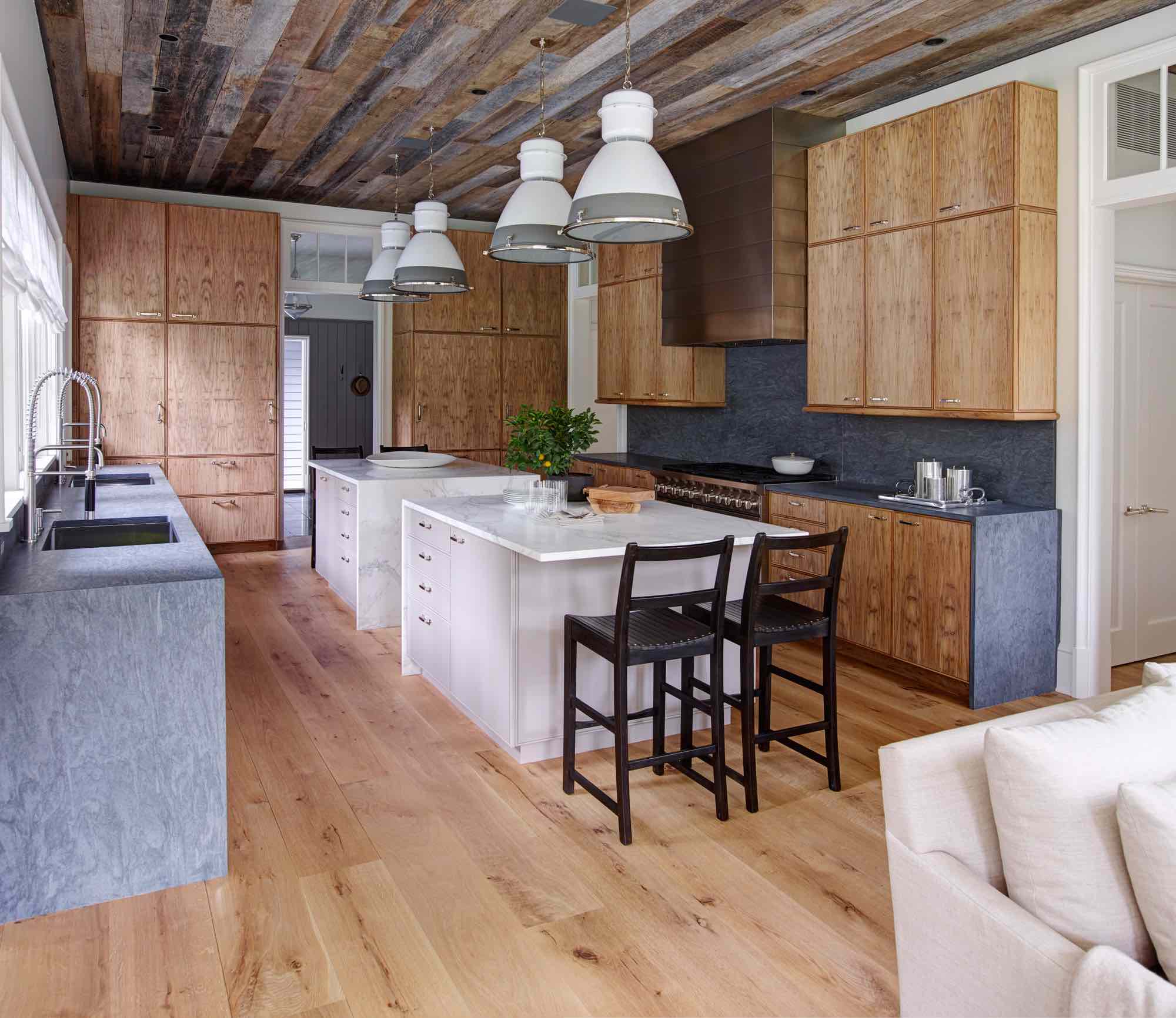 This image shows the knotty chestnut and soapstone kitchen cbinets with a pait of center Islands in a painted finish with Statuary Marble counter tops.  The kitchen A-M stoolwalnut barstools and Rover Factory industrial hanging pendant lights are by Ann-Morris Antiques. The range is by Wolf, and the ceiling is covered in reclaimed barn siding.  The cabinet Hardware is seen in a polished nickel finish   by Nanz Hardware.  Interior design by Carol Egan.