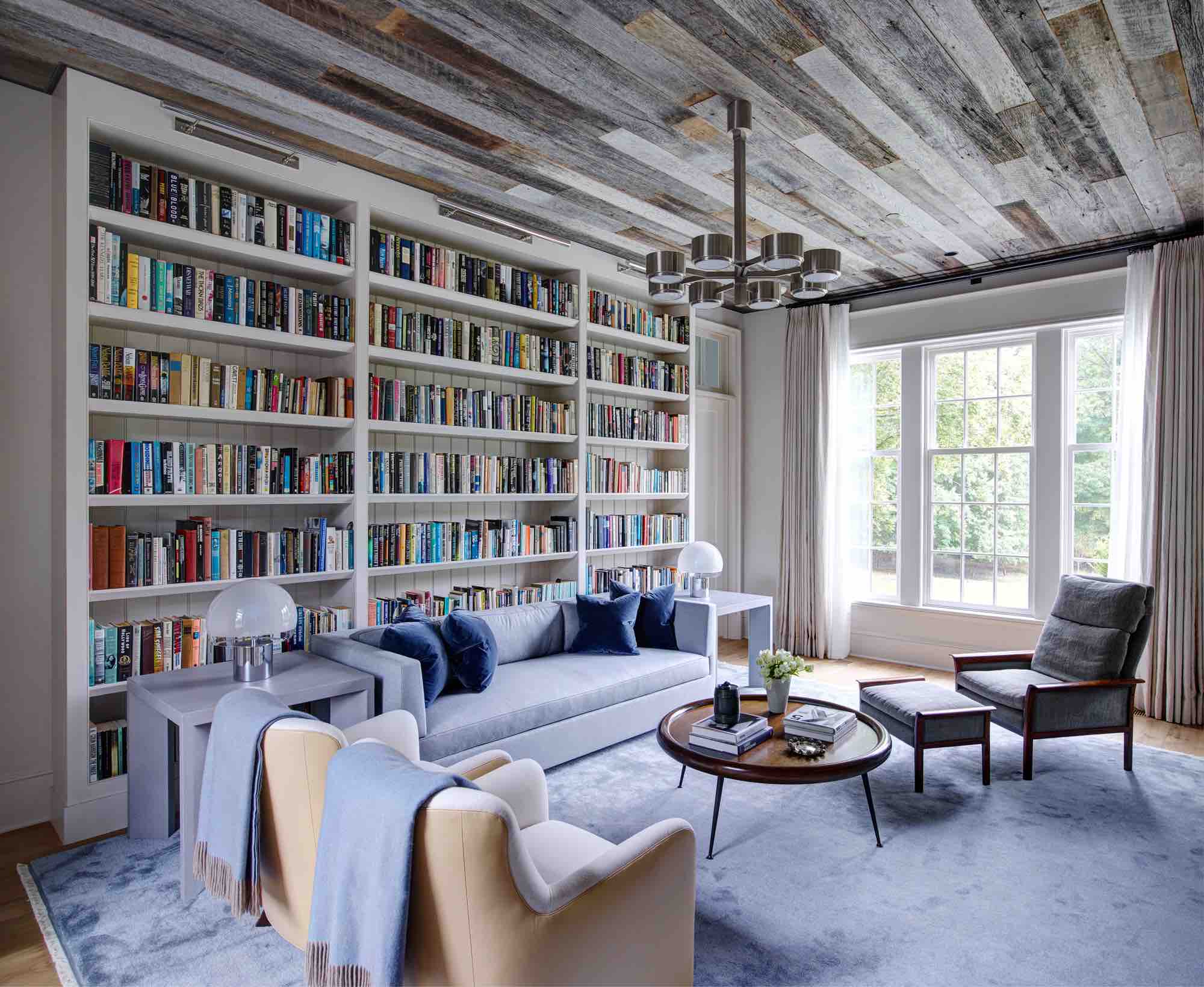 An image of the library in a Greenwich residence designed by Carol Egan.  The reclaimed barnwood ceiling is contrasted with painted millwork that encases the clients library.  There is a pale blue Nepalese silver carpet which compliments the putty color painted millwork.  The custom St. Thomas sofa in wool flannel smoke fabric by Holland & Sherry and Norwegian Rosewood lounge chair & ottoman surrounds a round walnut coffee table by Robsjohn Gibbins.  Table lamps by Luigi Caccia Dominioni.