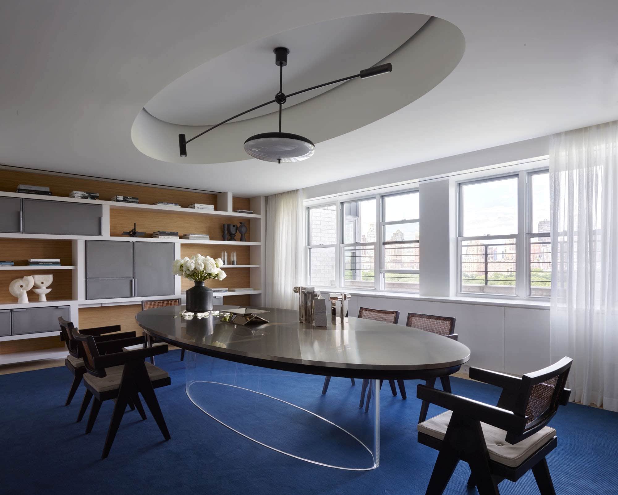 Shown in this image is a view of the library designed by Carol Egan with a custom commission table by Maria Pergay flanked by Chandigarh armchairs by Pierre Jeanneret, displaying Silver Candlesticks by Juan & Paloma Garrido and a ceramic vase by Georges Jouve.  A custom pendant design by Studio Carol Egan hangs above the dining table.  The curtains are in custom Cassiopea linen with integrated woven border by FJ Hakimian and the floor covering in custom rug by Sam Kasten.