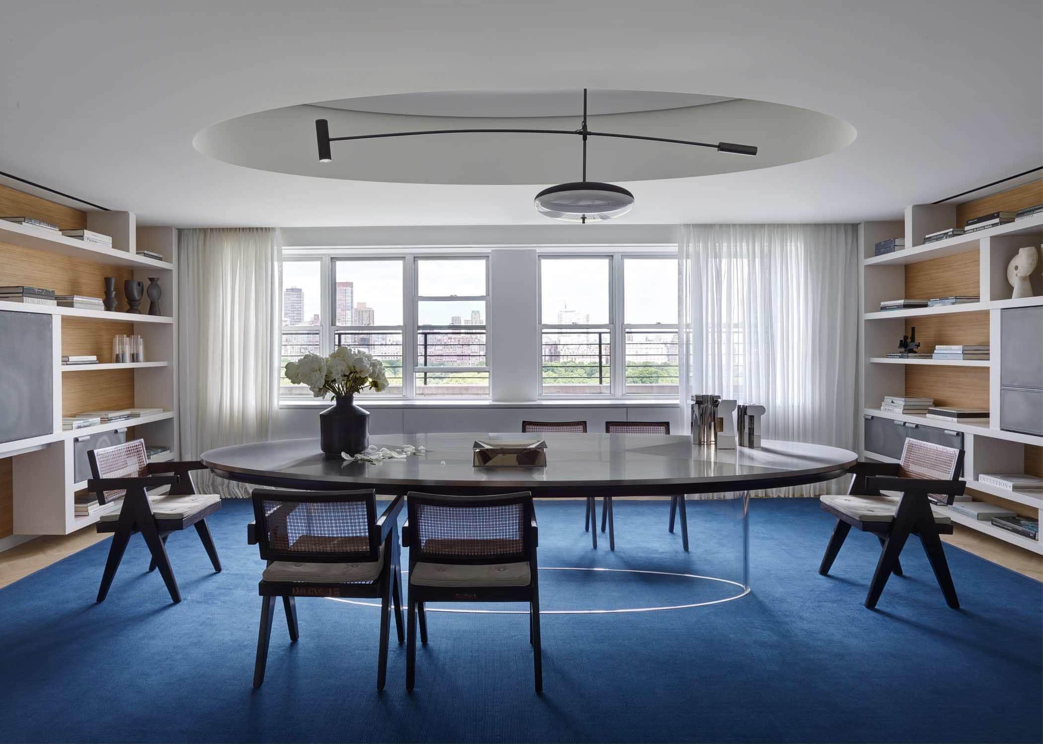 This photograph shows a library in a Fifth Avenue apartment.  The room consist of a custom commission table by Maria Pergay with Chandigarh armchairs by Pierre Jeanneret on a custom rug by Sam Kasten.  It is accessorized with silver candlesticks by Juan & Paloma Garrido and a ceramic vase by Georges Jouve and lit by a custom pendant design by Studio Carol Egan.  The curtains are in custom Cassiopea linen with integrated woven border by FJ Hakimian.