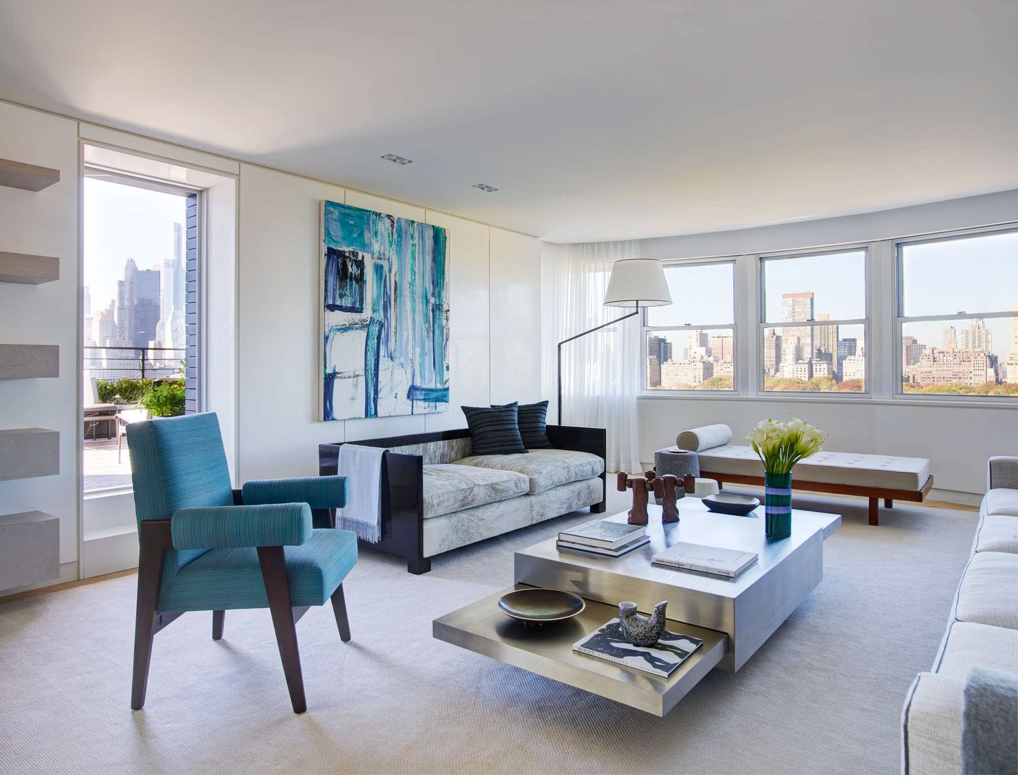 This is an image of a living room designed by Carol Egan.  In the middle of the room is a Tavolo 2000 coffee table by Gabriella Crespi in stainless Steel flanked by a1930s Swedish art deco sofa in custom grey charcoal boucle fabric by Sam Kasten, a Simple daybed by Charlotte Perriand in hand-woven fabric by Sam Kasten, a custom square arm sofa design by Studio Carol Egan, and armchair by Pierre Jeanneret.  A painting by Louise Fishman hangs on the wall beside a floor lamp by Angelo Lelli.  The curtains are in custom Cassiopea linen with integrated woven border by FJ Hakimian and the area rug is double sided from the summer/winter collection by Sam Kasten.