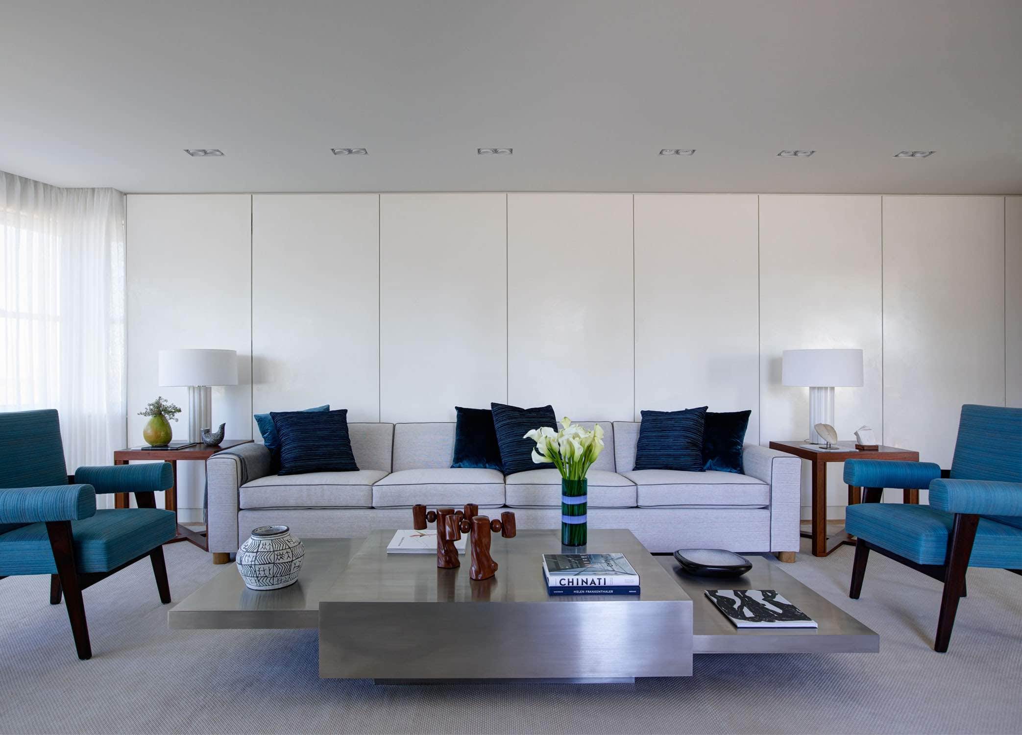 This image shows a living room designed by Carol Egan in a Fifth Avenue Apartment overlooking Central park in New York City.   A Stainless steel Tavolo 2000 coffee table by Gabriella Crespi can be seen in front of a custom square arm sofa design by Studio Carol Egan in a white and grey woven fabric by Toynie Seller.  Upholstered in blue are a pair of Adovcat armchairs by Pierre Jeanneret flanking the sofa.  A pair of Ephese white marble table lamps by Alexandre Biaggi sits on top of wooden sofa end tableside against a panele and plastered wall.