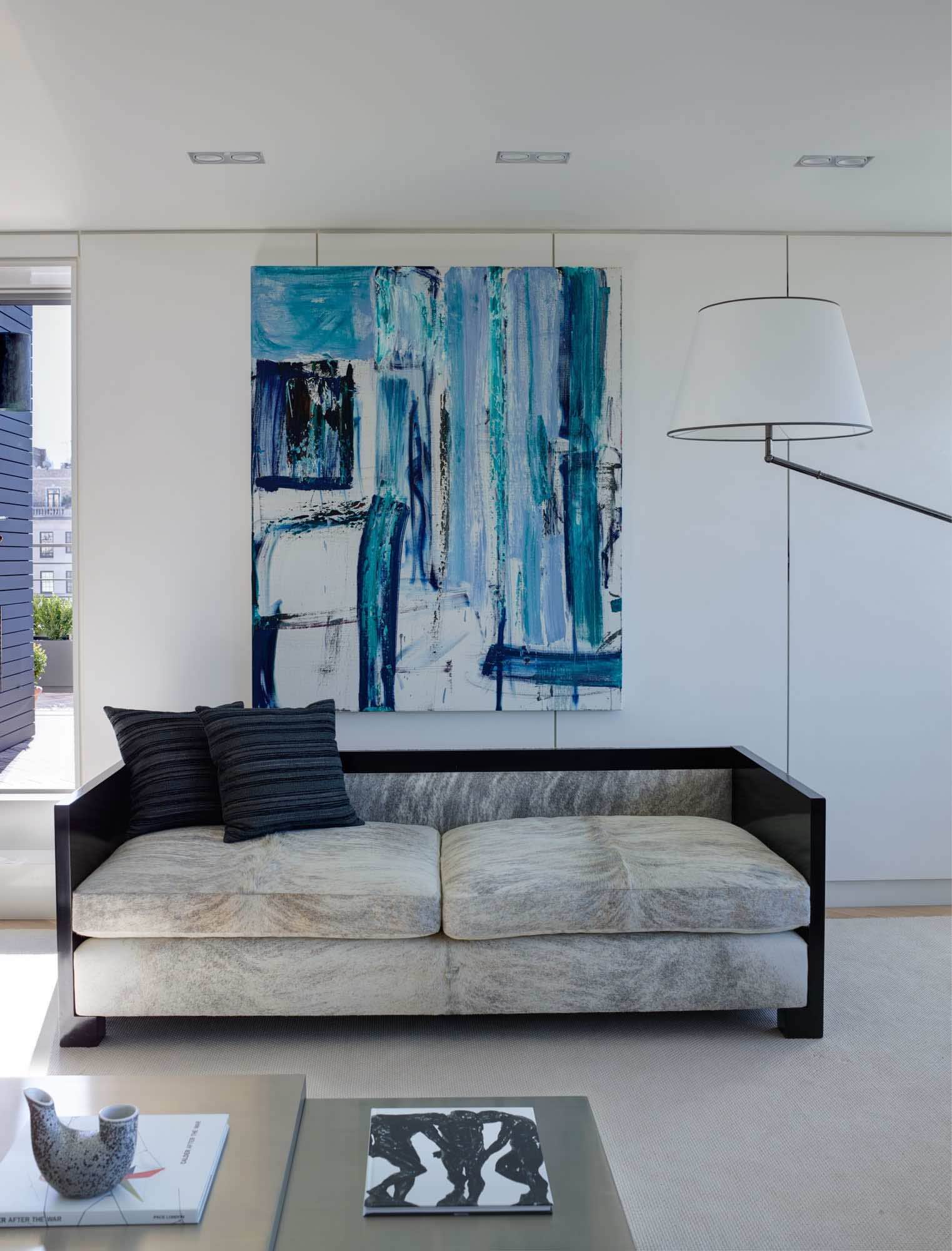 Shown in this image is a view of the living room designed by Carol Egan in a Fifth Avenue Apartment which overlooks Central Park in New York City.  A painting by Louise Fishman hangs behind a 1930s Swedish art deco sofa with ebonized wooden frame and grey cowskin upholstered seat and a vintage floor lamp by Angelo Lelli.