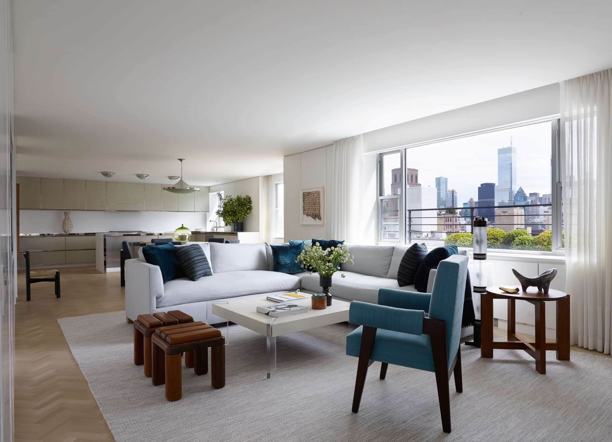 Shown in this image is a family room and kitchen in a Fifth Avenue Apartment designed by Carol Egan and overlooking Central Park in New York City.   An L-shaped sofa is upholstered in a grey wool fabric, Almofada wooden stools by Zanini de Zanine Caldas and an armchair by Pierre Jeanneret flanks a coffee table by Vladimir Kagan in white acrylic and Lucite base.  Next to the sofa is a swedish wooden side table in stained oak.  The curtains are white woven lined with an integrated woven border that aligned with the sill height around the room.