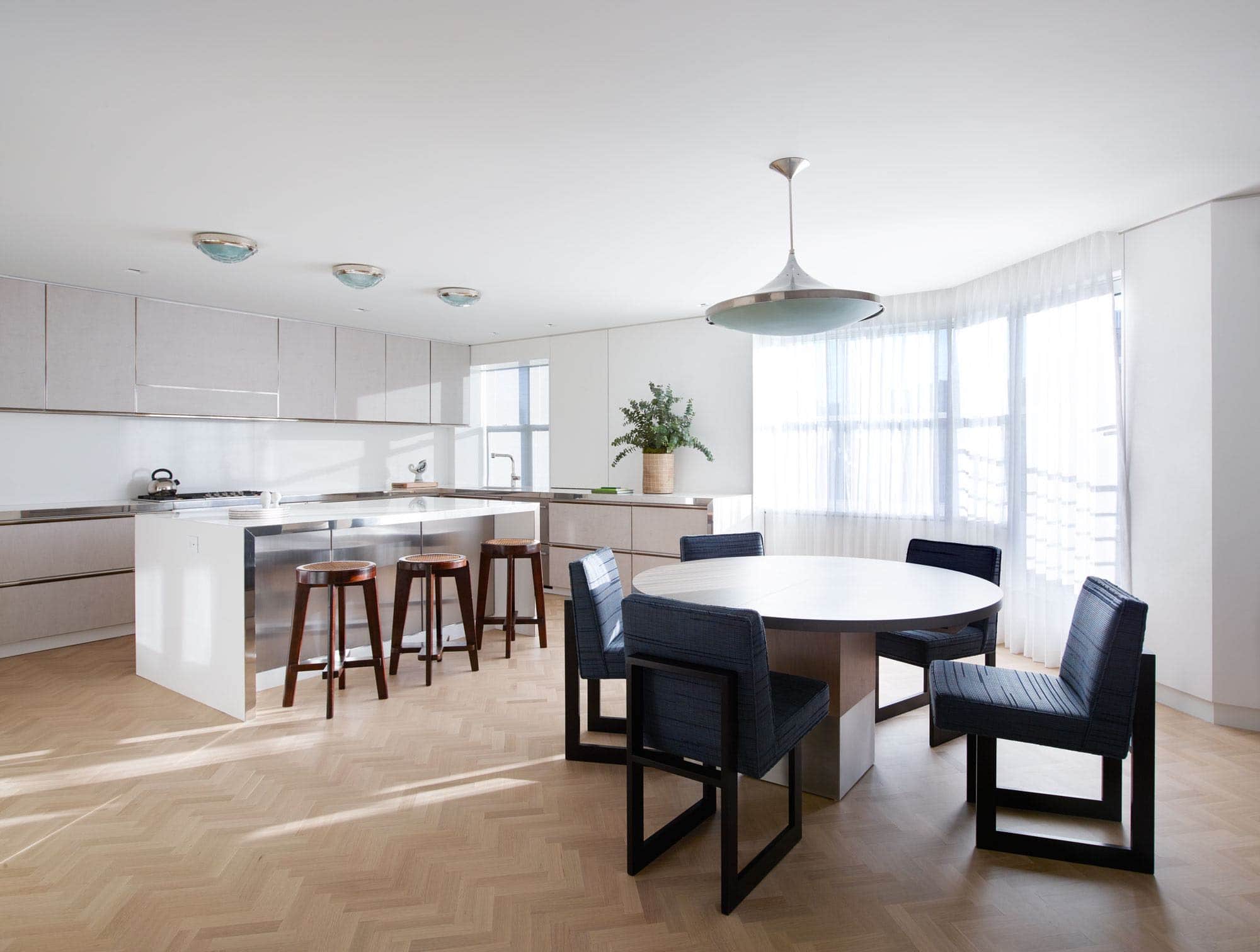 This photograph shows the view of a kitchen and breakfast room in an apartment on Fifth Avenue which overlooks Central Park in New York city designed by Carol Egan.  A Chandelier by Fontana Arte made from stainless steel and cast glass hangs above a custom smoked oak dining table by Van Rossum with upholstered armless dining chairs by Vladimir Kagan in a custom wool and woven leather blue fabric.