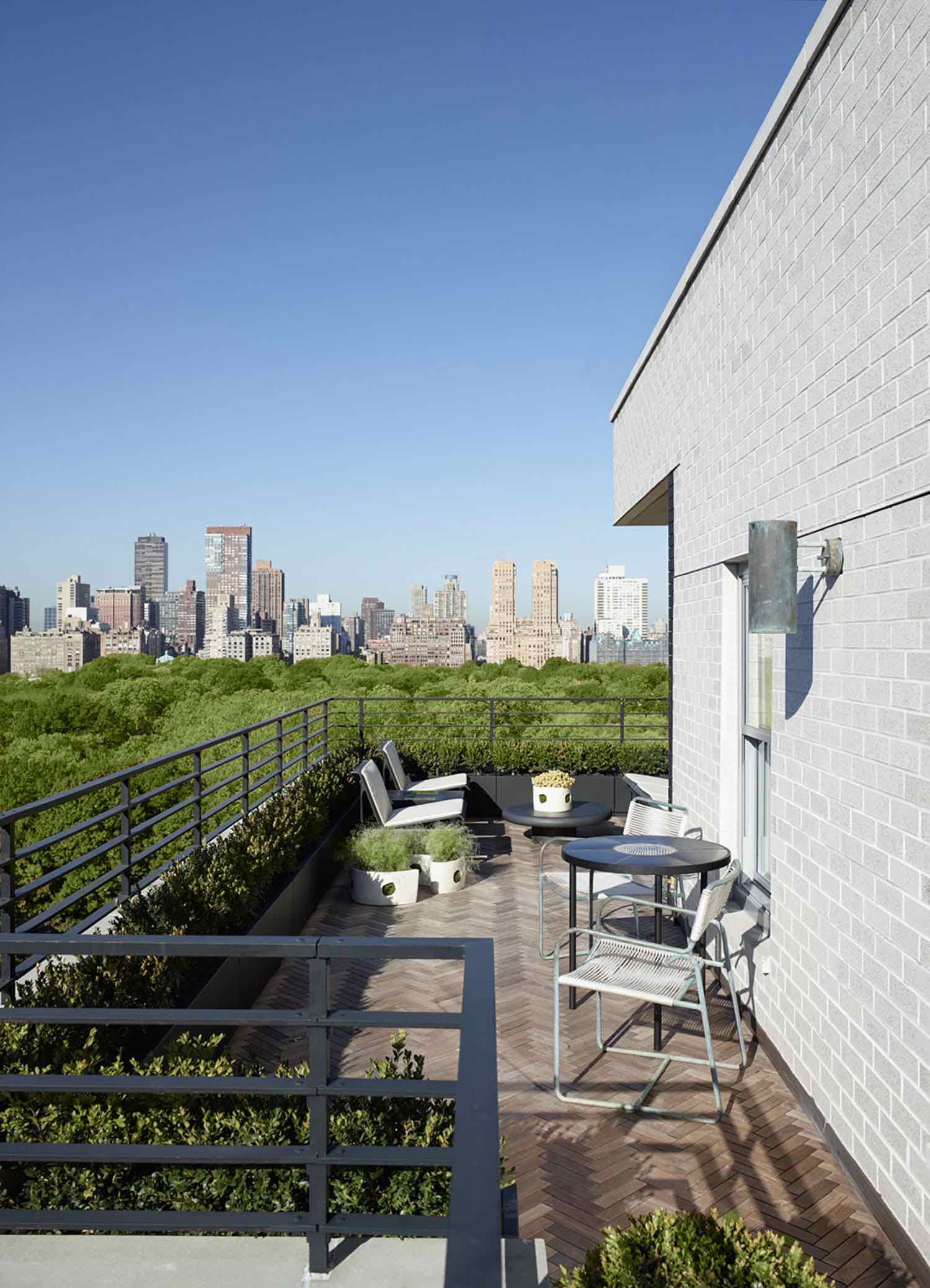 This image shows a terrace designed by Carol Egan in a fifth Avenue Apartment which overlooks Central Park in New York City.  A ceramic dining table by Marianne Vissiere with armchairs by Walter Lamb are seen in the foreground sitting on a custom teak herringbone deck.   Oxidized copper wall lights by Hans Agne Jakobsson Are installed on the exterior walls.