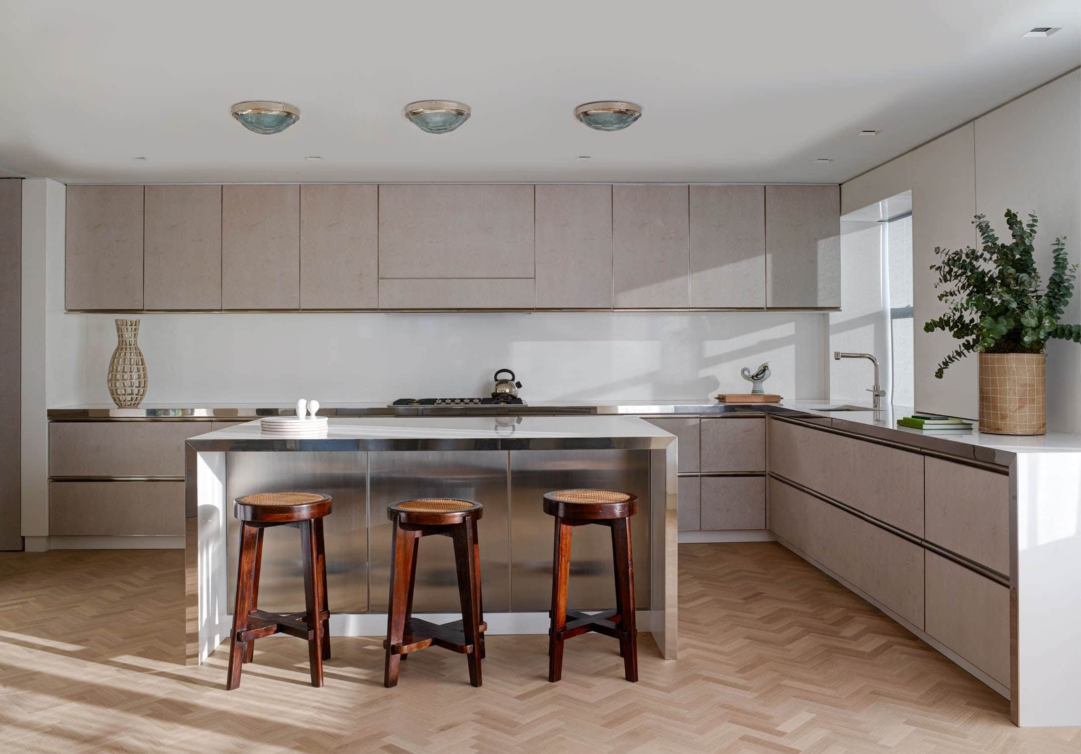 Designed by Carol Egan, this image is of a kitchen with three 1960s ceiling lights by Fontana Arte above a kitchen island in Swedish ice birch over and hone marble with three vintage barstools by Pierre Jeanneret.  Swedish ice birch cabinets with custom designed integrated metal door pull make up the L shaped Kitchen millwork.  A bucket vessel by Kathy Erteman and a Spanish vase are displayed on the countertops.