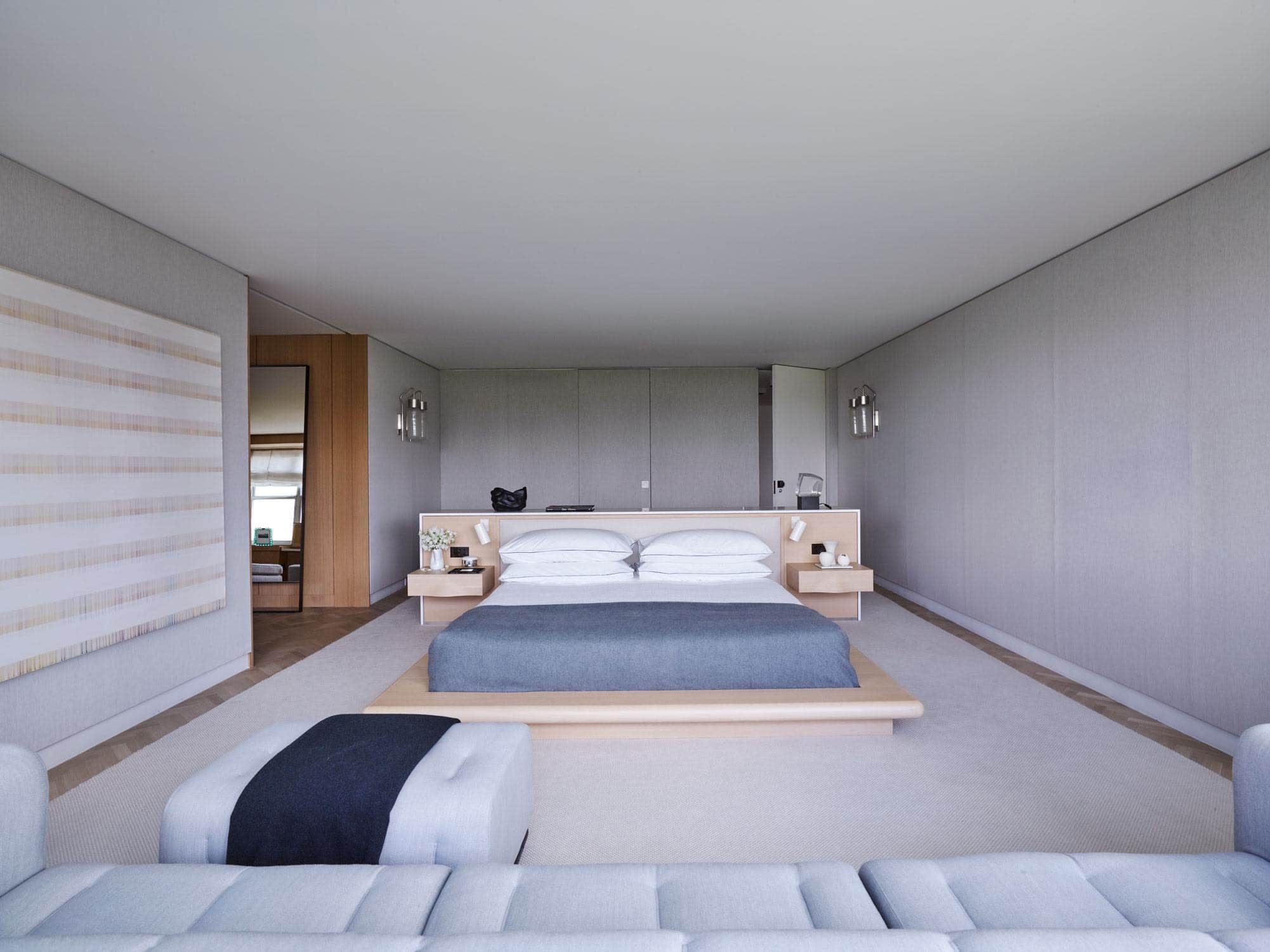 This photograph shows the master bedroom in a Fifth Avenue apartment designed by Carol Egan which overlooks Central Park in New York City.  With upholstered grey linen walls, the custom designed platform bed floats in the center of the room.  A vintage Scarpa sofa upholstered in grey flannel with ebonized mahogany frame sits in the window.