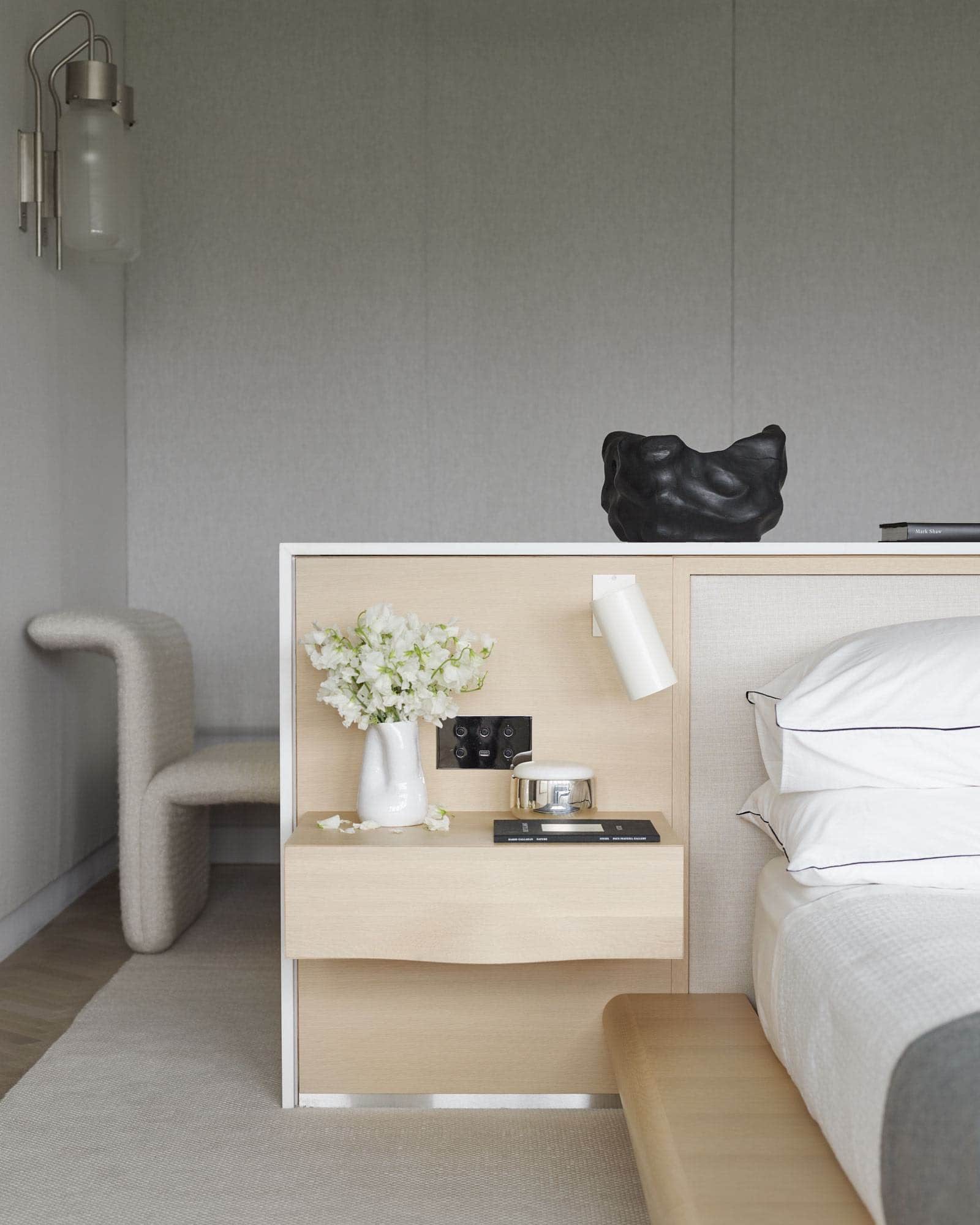 Shown in this image is a detail of the bedside table integrated into a platform bed design in the master bedroom of a fifth avenue apartment designed by Carol Egan.  The bed is comprised of a mill oak undulating Cabinet, capped in white marble with a honed finish and fitted with a linen upholstered headboard and an oak platform and bedside tables.  Bidone wall lamps by Luigi Caccia Dominioni are installed on the linen walls for wall light.  The area rug is a woven natural linen.