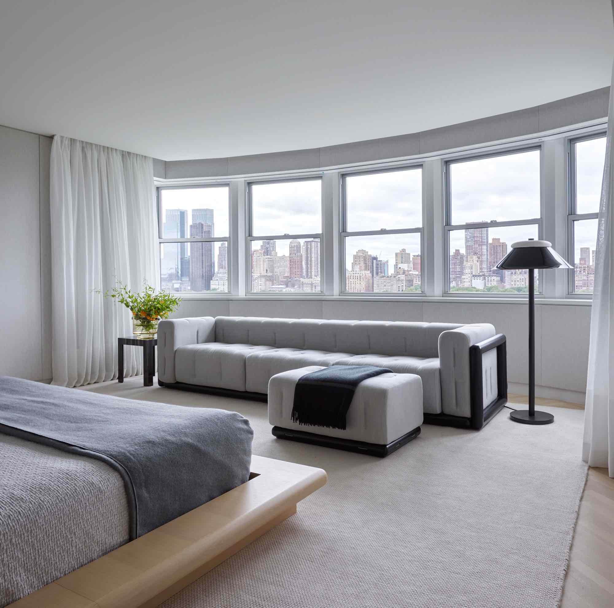 This image shows a view of the sitting area in a master bedroom designed by Carol Egan in a Fifth Avenue Apartment which overlooks Central park in New York City.   A vintage Cornaro 220 sofa by Carlo Scarpa with ebonized wood frame is upholstered in a Loro Piana wool flannel in grey.  Next to the sofa is a floor lamp by Eric Schmitt in blackened bronze and alabaster.   On the left side of the sofa is a side table by Alexander Zhikulin in steel that has been patinated to look like strae marquetry after Jean Michel Frank table design.  The curtains are white woven lined with an integrated woven border that aligned with the sill height around the room.