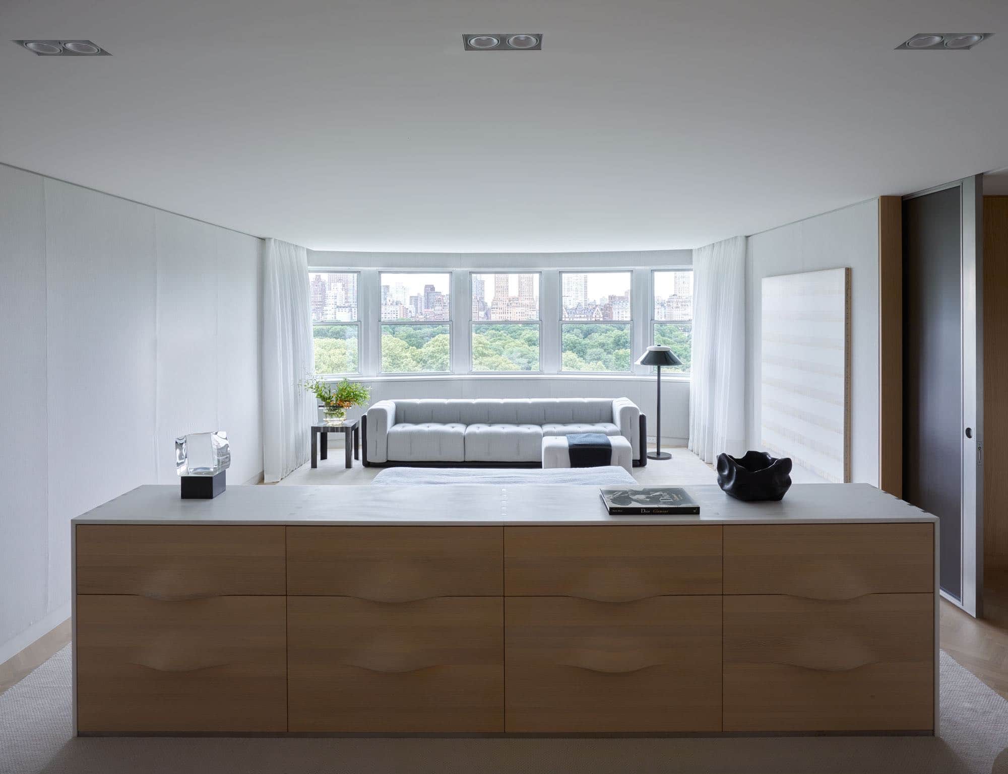 This photograph shows a master bedroom designed by Carol Egan in an apartment on Fifth Avenue in New York City which overlooks Central Park.  A headboard design which is integrated with a cabinet with undulating drawer fronts in Oak by Studio Carol Egan divides the room upon arrival.  the cabinet is two sided, a function cabinet as one side and an upholstered platform bed from the other side.  Displayed on the marble top of the cabinet is a Loukoum table lamp by Christophe Come in Cast Glass and blackened metal.   A Scarpa Sofa sits under the window with an "Emily" floor lamp by Eric Schmitt.