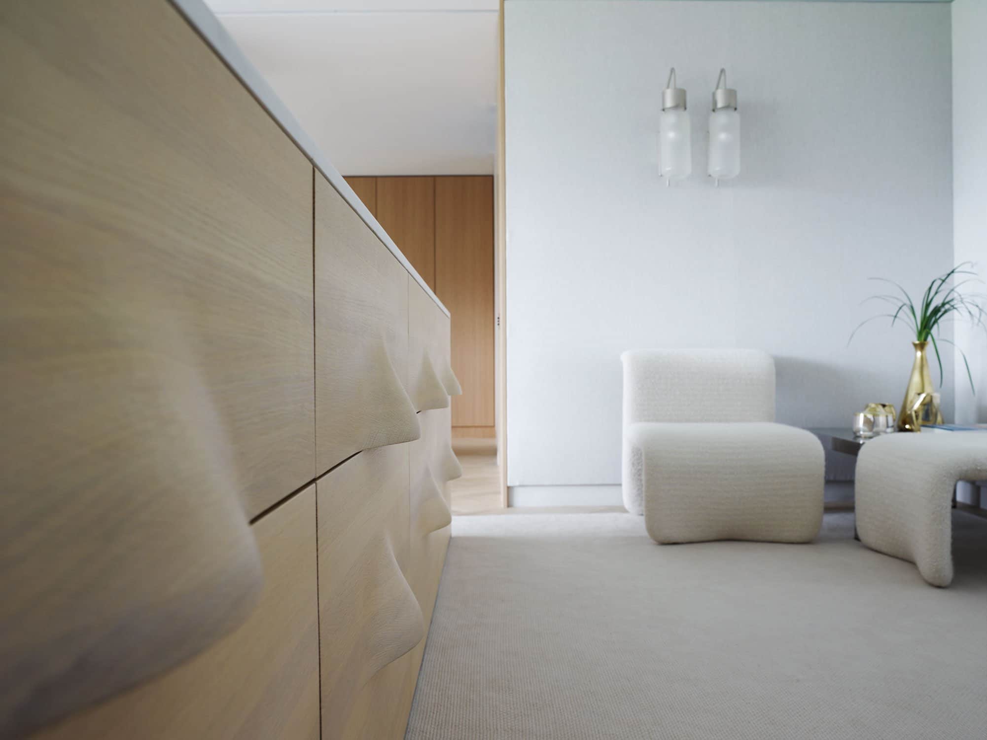 Designed by Carol Egan, this image shows the view as you enter the master bedroom in an apartment on Fifth Avenue in New York city which overlooks Central Park.  A detailed view of a cabinet headboard can be seen with its undulating pattern in milled and laminated oak.  The walls of the room are upholstered in grey linen and a pair of Bidone wall lamps by Luigi Caccia Dominioni hang above a seating area.