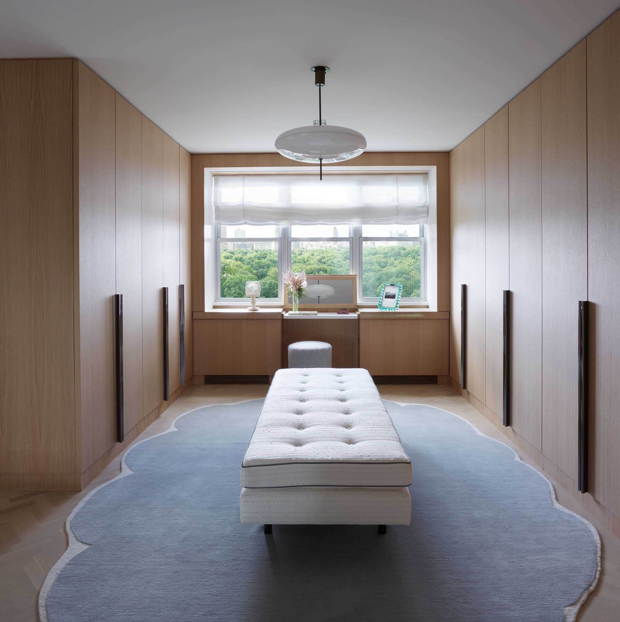 Shown in this image is a Master dressing room in an apartment on Fifth Avenue in New York which overlooks central Park.  The view of the park is framed in custom oak millwork with a flip top vanity build into the window seat. A cloud shape Area rug designed by Francois-Xavier & Claude Lalanne sits on the herringbone oak floor.  A vintage 2065 pendant light by Gino Sarfatti hangs from the ceiling above a custom daybed.  A 1940s alabaster table lamp from Rose Uniacke is in the window.