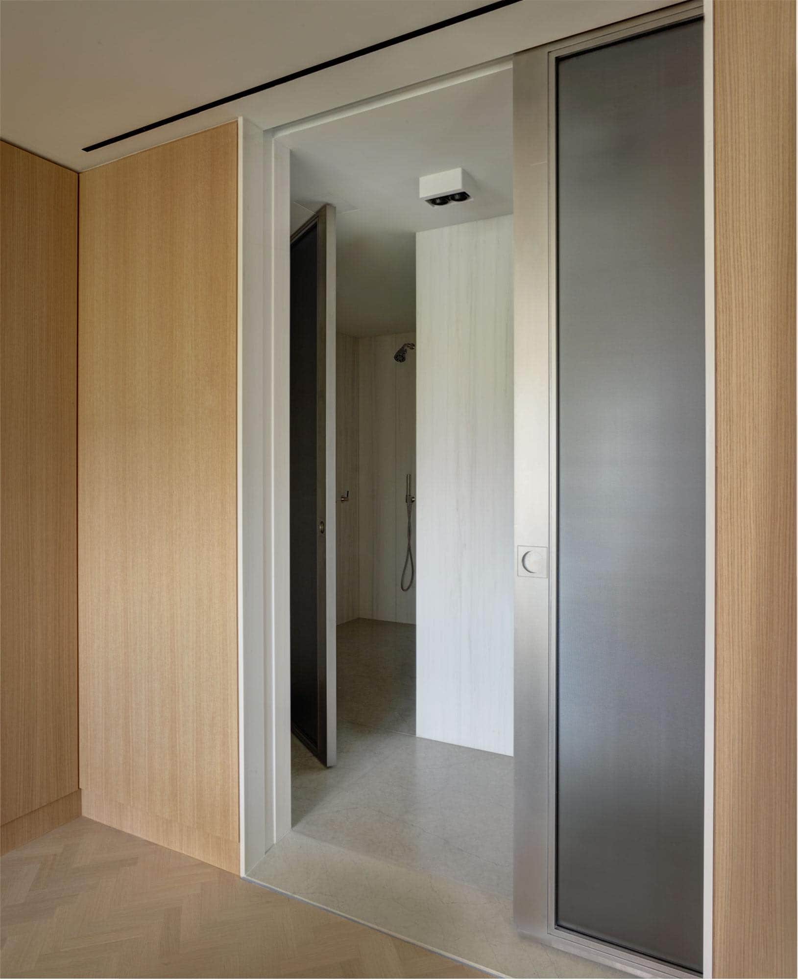 Shown in this image is a view into the master bathroom in an apartment on Fifth Avenue in New York.  Oak wall panels frame a pocket door which houses a steel and ribbed glass sliding door which leads to the Slab Marble paneled master bathroom.