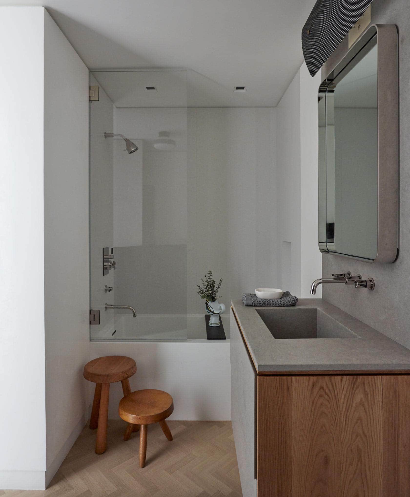 The image shows a guest bathroom designed by Carol Egan with a perforated rectangular wall lamp by Pierre Guariche above a mirror by Gio Ponti and a square tailor washbasin integrated in a plana shelf and wall made of Sinai stone.  Hung on the stone wall is a Gio Ponti Mirror and the wooden milking stools are by Charlotte Perriand.