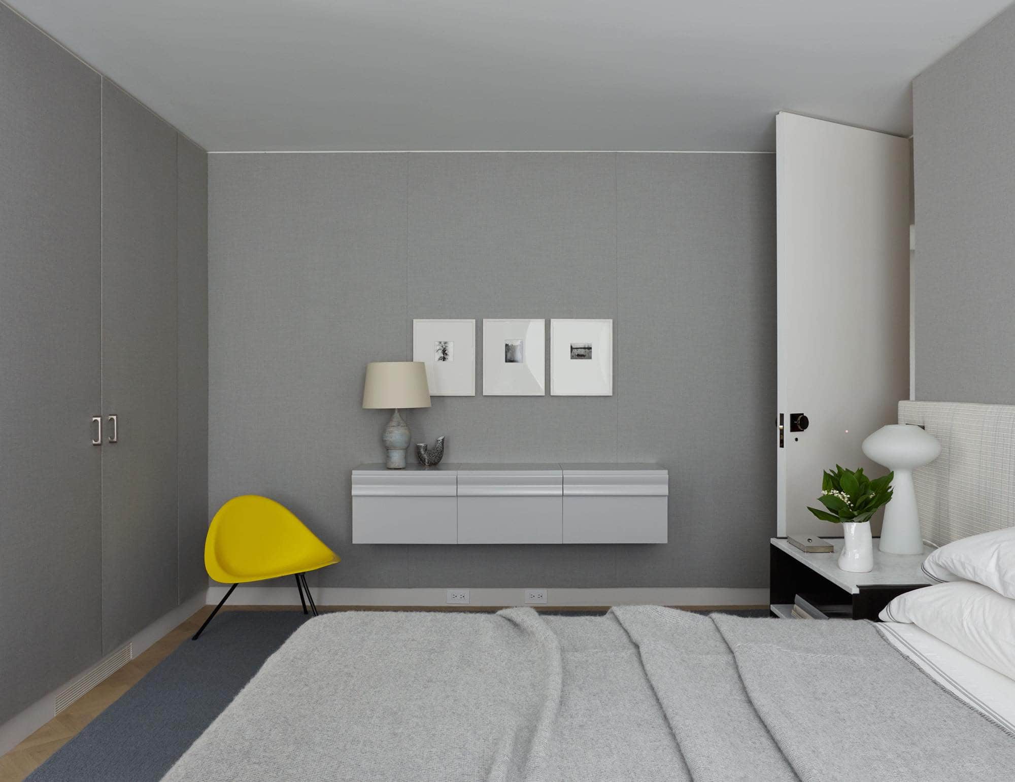 This image shows a guest bedroom designed by Carol Egan in a residence on Fifth Avenue in New York.  With a custom bed upholstered in a suede and wool woven leather and installed in a wall-to-wall condition. A blanket in Chedi natural grey silk fabric by Soie de Lune is draped over the bed.  The bedside tables are metal and marble and are set in front of the upholstered headboard side panel.  The walls of the room are upholstered in grey wool flannel and a cantilevered lacquered grey cabinet hangs on the upholstered wall.  Adjacent to it is a yellow aluminum tripod chair by Poul Kjaerholm.  A blue flecked ceramic table lamp by Jacques Blin sits on the cabinet.