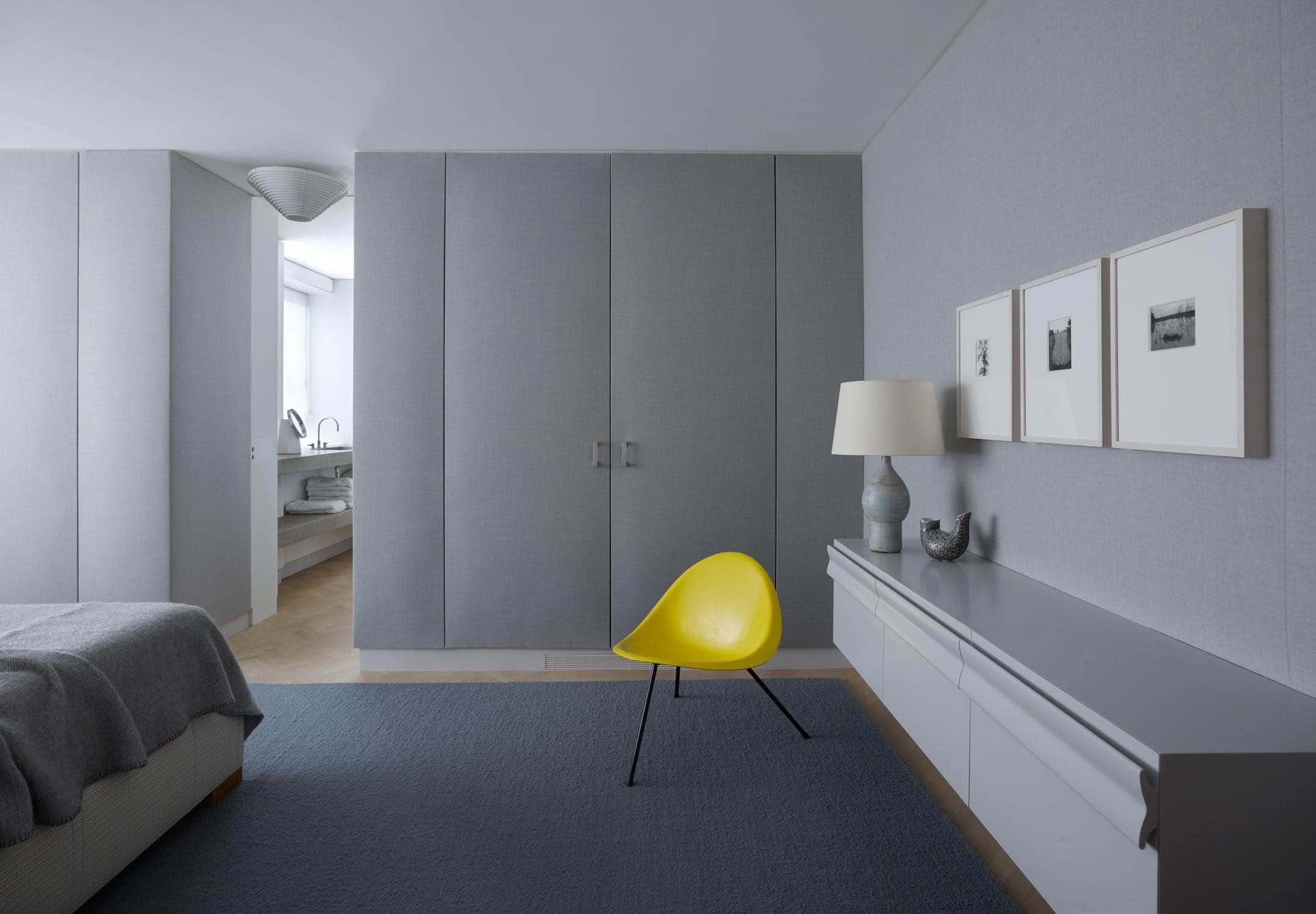 Designed by Carol Egan, this guest bedroom is located in a residence on Fifth Avenue in New York which overlooks Central Park. The bedroom is upholstered in Grey wool flannel.  A custom designed grey lacquered cabinet is cantilevered from the bedroom wall with a yellow aluminum tripod chair by Poul Kjaerholm contrasting the monochrome.  A grey chunky boucle area rug contrasts the natural herringbone oak flooring.