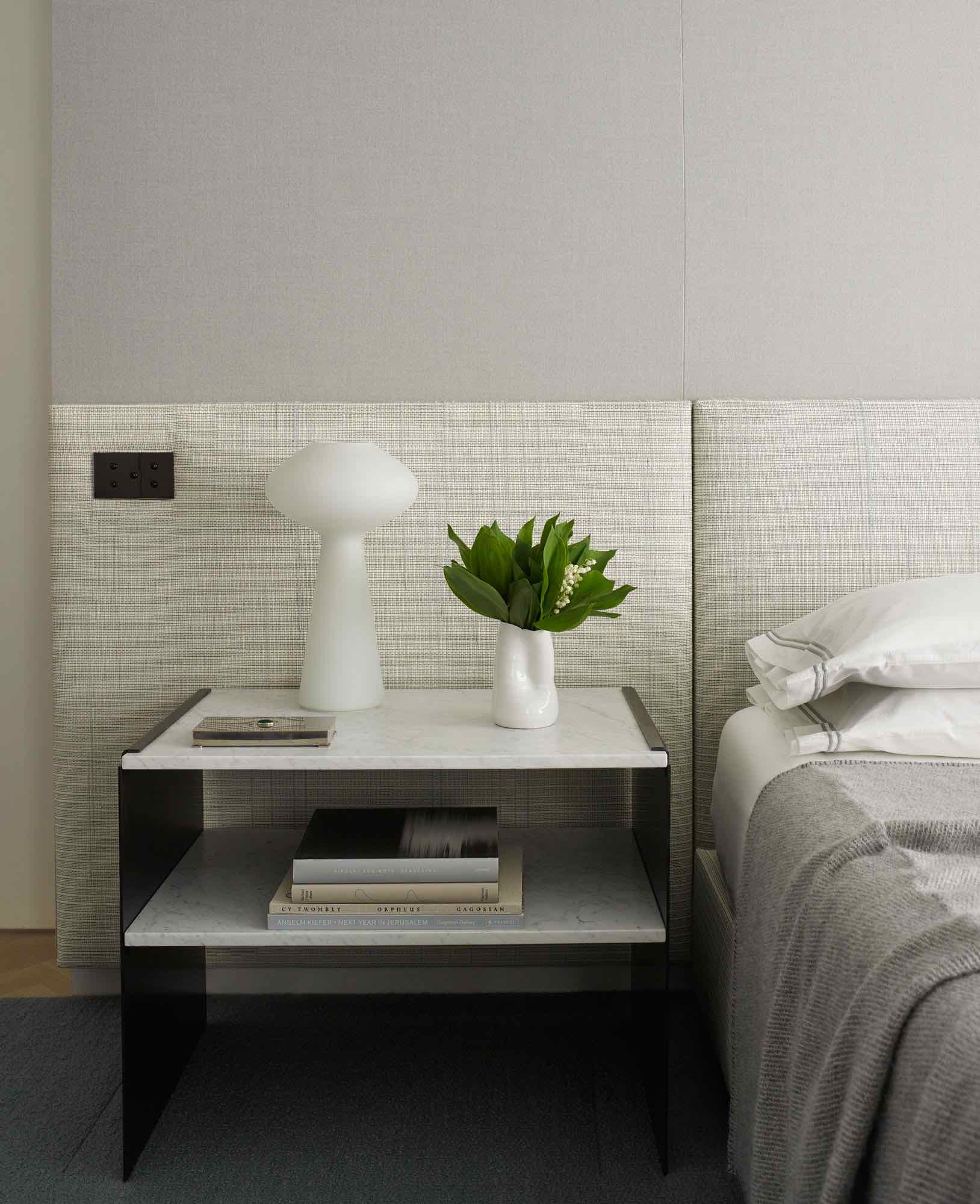 This photograph shows a custom bed design by Studio Carol Egan in a guest bedroom in a residence on Fifth Avenue in New York.  A marble and blacked bronze canal side table flanks the bed. with a white glass mushroom table lamp by Lisa Johansson-Pape.  Wall Upholstery in Elizabeth St. bespoke twill fabric by Holland & Sherry.  There is a ceramic cup with a bouquet of Lilly of the valley on the bedside tabletop.