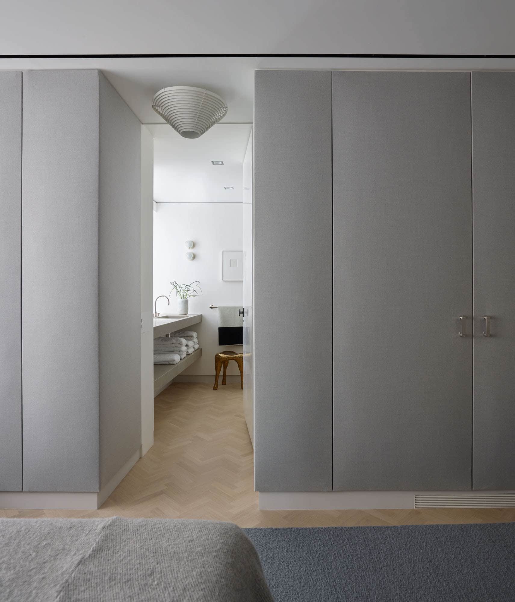 Shown in this image is a view of guest bedroom and bathroom in a residence on Fifth Avenue in New York.  The walls of the room are upholstered in a grey wool flannel and the floors are a natural oak herringbone pattern.  An A226B ceiling light by Alvar Aalto hangs on the ceiling.  In the bathroom, glass Joe Colombo Fresnel lights are stacked on the wall by the stone sink and a Hex stool by The Haas Brothers is set next to the sink.