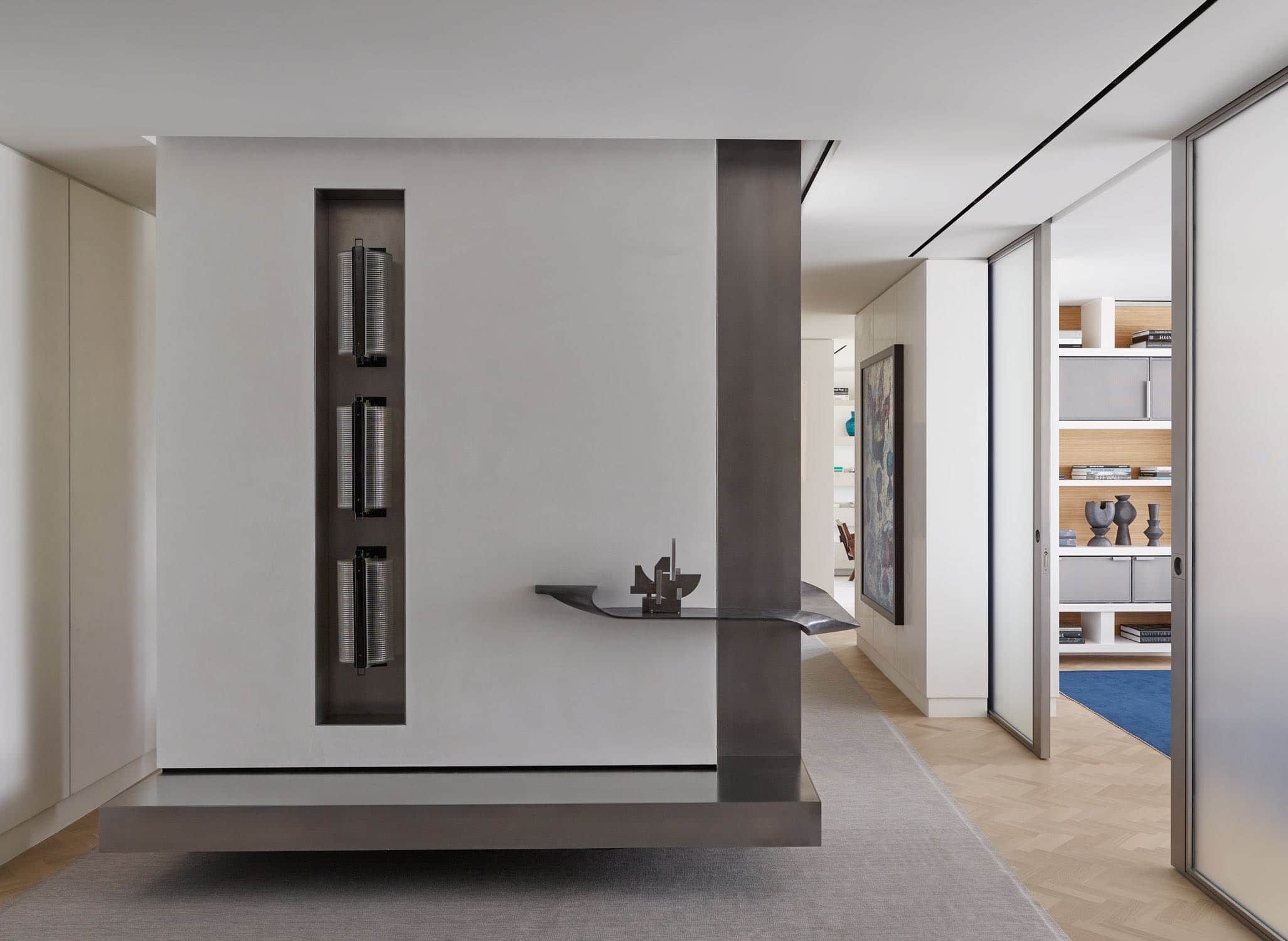 A photograph of a gallery designed by Carol Egan in a residence on Fifth Avenue in New York City.  Walls are plastered with insets of steel panels shown with a custom wall hung shelf - leather wrapped by Studio Carol Egan displaying a sculpture by Marino Di Teana.  Three Vintage Stilnovo Cast Glass wall sconces are recessed in a steel niche and the custom grey heathered wool area rug is bordered by a herringbone oak floor.