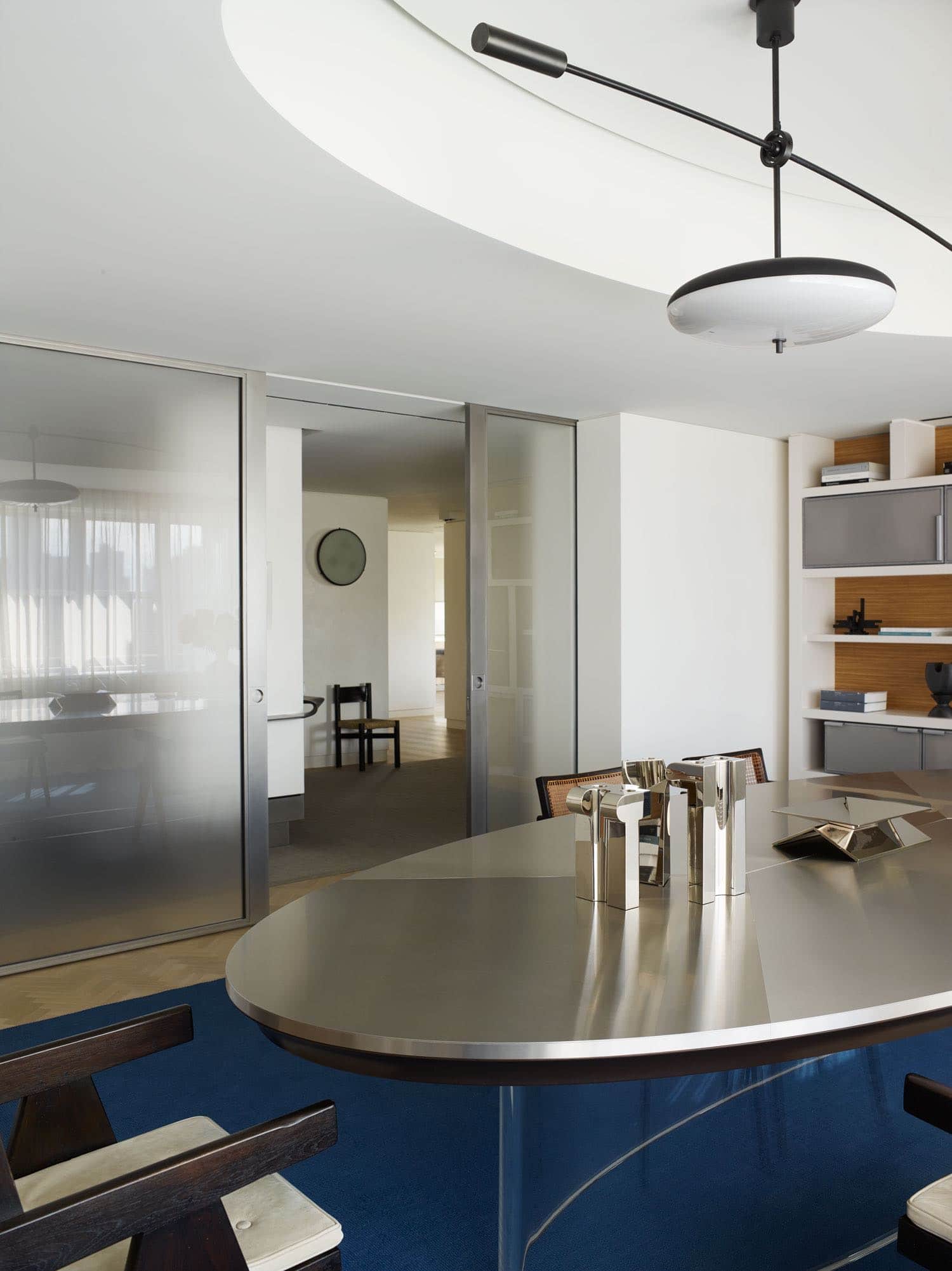 Designed by Carol Egan, this image is of a library which overlooks Central Park in New York City.  With a custom commissioned Steel and Lucite dining table by Maria Pergay flanked by Chandigarh armchairs by Pierre Jeanneret on a custom silk Ultramarine Blue area rug by Sam Kasten.  Displayed on the table are silver candlesticks by Juan & Paloma Garrido, silversmiths from Spain.  A custom pendant sculptural light fixture in blackened metal and acrylic hangs above the table.  Custom Steel and ribbed glass pocket doors can be seen in the background.