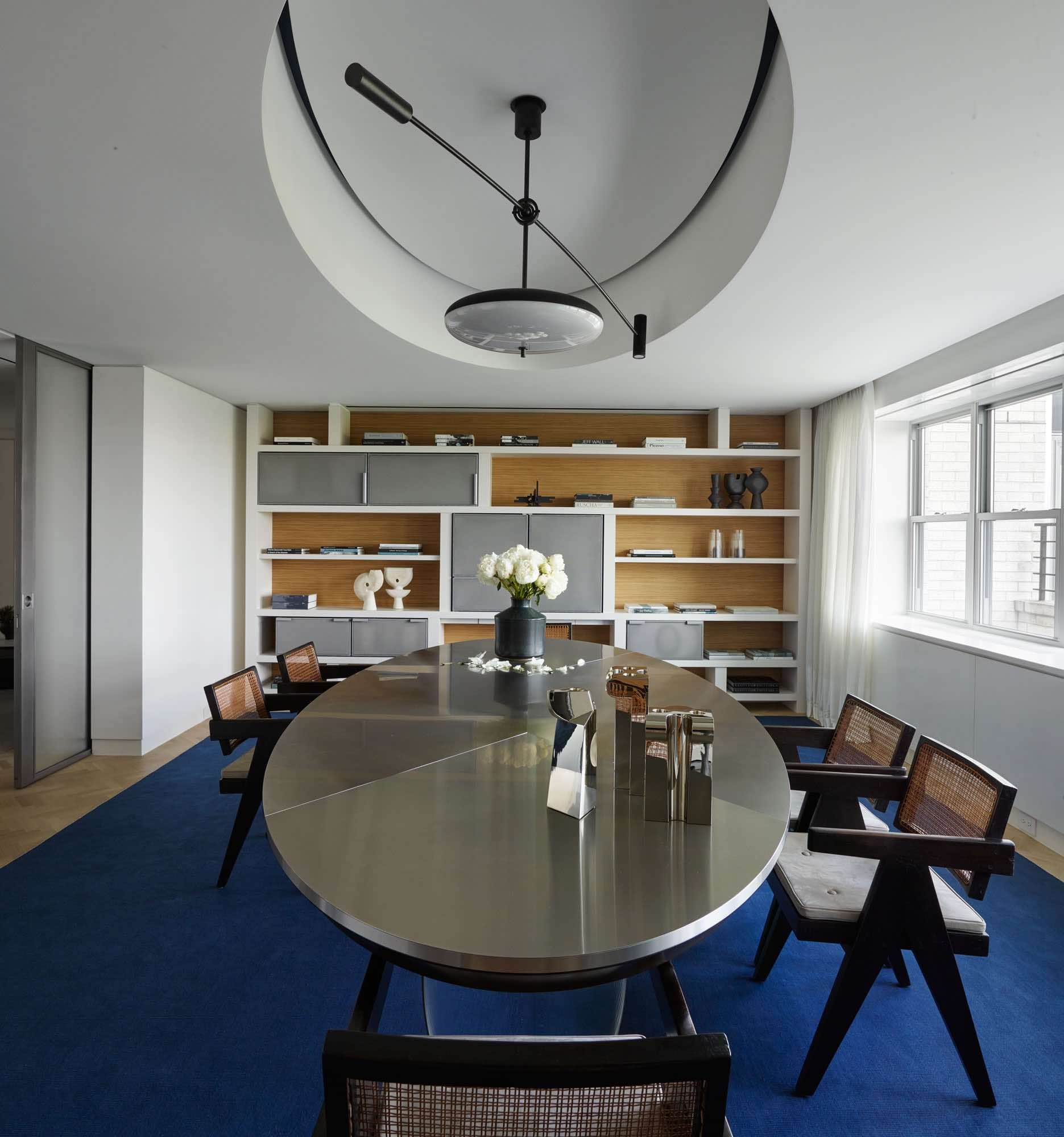 This image shows a library designed by Carol Egan.  A custom commission table by Maria Pergay sits in the middle of the room surrounded by Chandigarh armchairs by Pierre Jeanneret.  A custom pendant design by Studio Carol Egan hangs above the dining table showcasing silver Candlesticks by Juan & Paloma Garrido and a ceramic vase by Georges Jouve.  The curtains are in custom Cassiopea linen with integrated woven border by FJ Hakimian and the area rug in the room is a custom silk ultramarine color made by Sam Kasten, American handweaver.