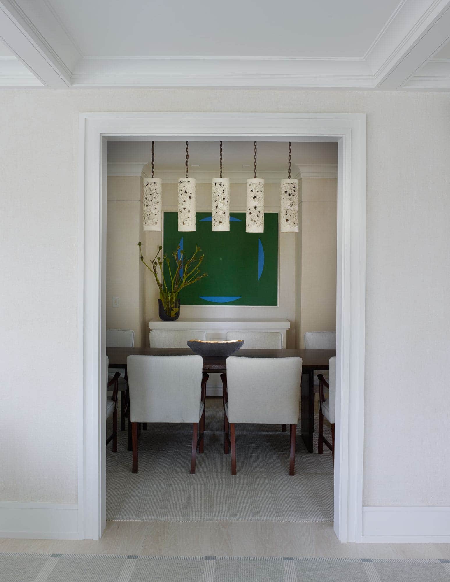 This image shows a dining room designed by Carol Egan with artwork by Jorge Fick and Garden Grid area rug by Elizabeth Eakins.  There are 5 Vintage cylindrical pendant lights in porous pottery hanging above the Japanese wooden dining table by Tucker Robbins that is flanked by dining chairs by Ole Wanscher.  There is a paper buffet by Job Smeets with a ceramic and bronze bowl by Cristina Salusti, Combed Gesso wall treatment by Boyd & Reathe and Hill bronze vase by Eric Schmitt.