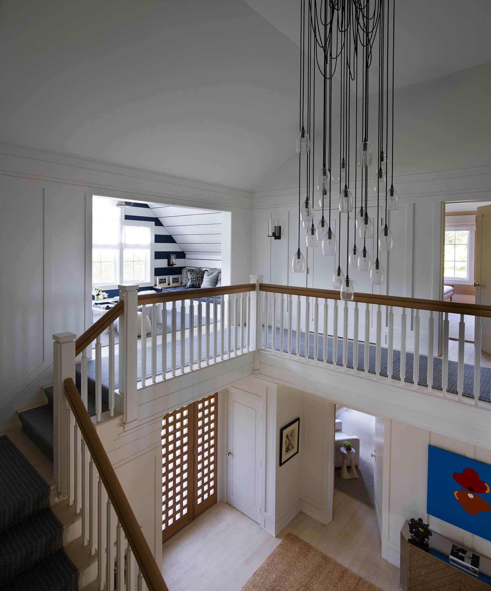 This image is a view up the stairway to the second-floor hallway in the Bridgehampton home designed by Studio Carol Egan.  A Scripted pendant chandelier by Alison Berger hangs from the ceiling in blackened Bronze and engraved glass pendants that are contrasted against the white shaker panels of the 2nd Floor stair hallway.  There is a custom woven runner carpet on the stairway - Deep blue basket weave by Elizabeth Eakins.  You can see a nook at the top of the stair with built in banquettes for playing chess and backgammon.