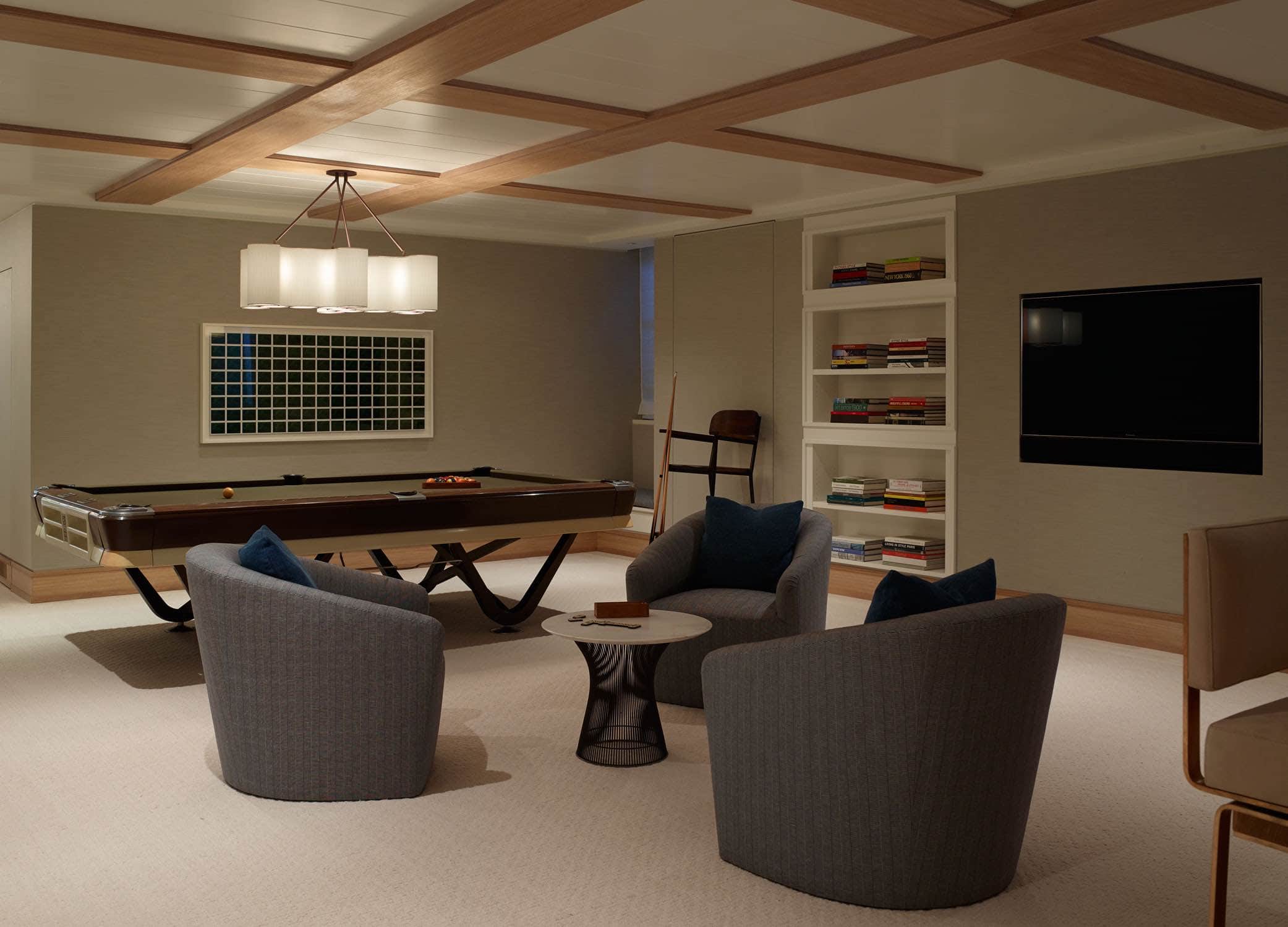 This image shows a games room designed by Carol Egan with a vintage pool table.  The pool table is lit by a ""Linear Cumulus"" pendant fixture by Ted Abramczyk.  The walls are upholstered in a taupe fabric and the ceiling is accented with a shaker oak and painted white wooden grid. Three swivel armchairs that are upholstered in a black and white woven fabric sit on a Custom ramie & wool ""Cuddles ll"" flat weave area rug by Mitchell Denberg.