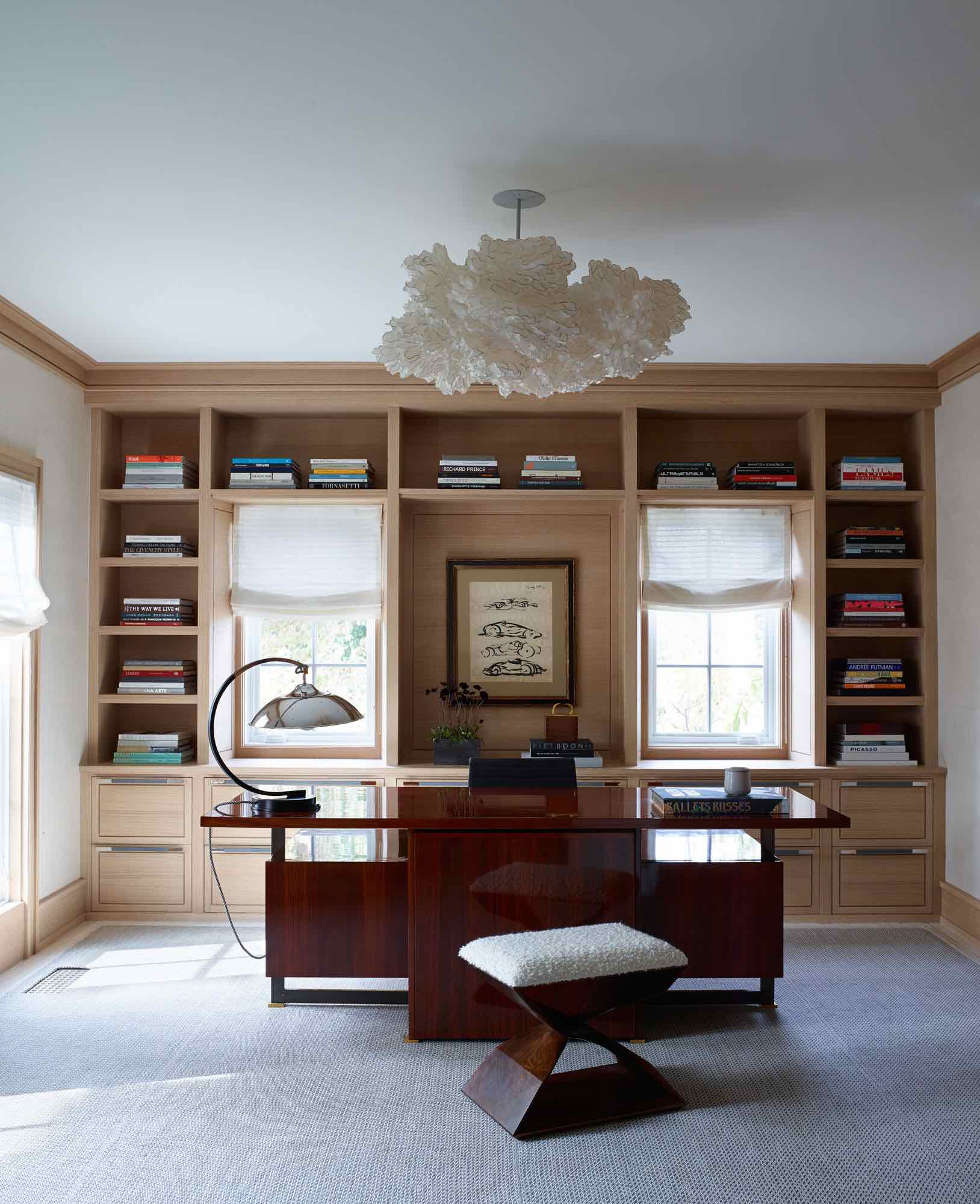 This image shows a library with custom designed oak millwork bookcases in this Bridgehampton home designed by Studio Carol Egan.  There is a ceiling light fixture called the "Cloud" by Ayala Serfaty that hangs over a Leleu Desk in Mahogany finish.  On the custom oak millwork bookcases are leather upholstered cabinet drawer pulls by Holland & Sherry.  In the foreground is an X bench in a walnut finish designed by Studio Carol Egan.  There is a custom woven area rug in the library by Elizabeth Eakins.