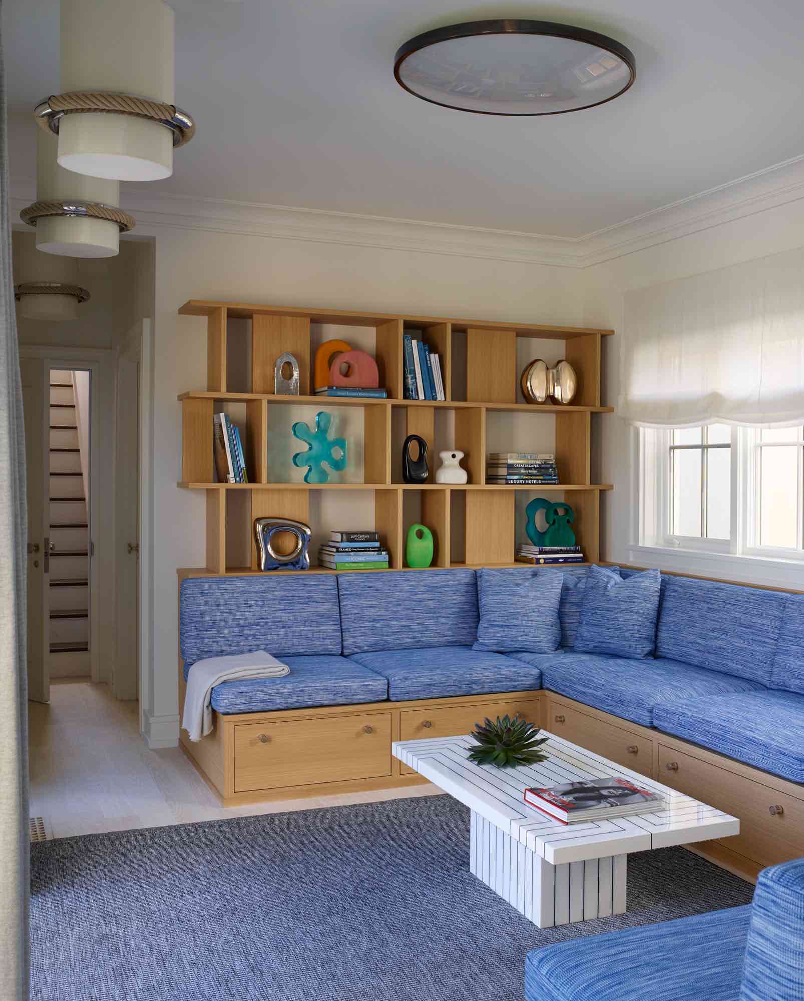This image shows a built-in bookcase and seating nook in white rift oak designed by Carol Egan.  The ceiling fixture is a flush mounted Vintage 1960 patinated metal circular ceiling light, and the adjoining hallway shows the sailor pendant lights by Thomas Boog.  There is a custom area rug by Sam Kasten along with custom handwoven acrylic fabric for sofa by Sam Kasten on the banquette.  Sculptures by Mia Fonssagrives are displayed in the bookcases and a Daytona 200 coffee table by Peter Dayton.