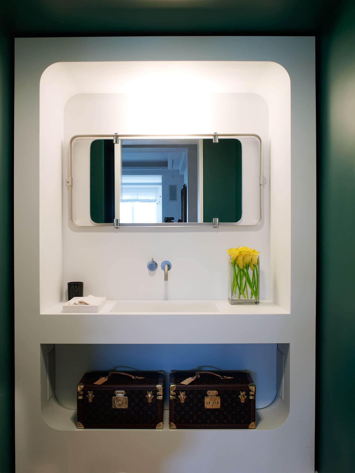 This image shows a powder room with a white Corian framework enclosing the sink and niche shelves below the sink.  The room is painted in a pantone modern green color contrasting the white Corian framed sink wall.  Hung on the Corian wall is a modernist mirror with steel frame and vintage train travel cases by Louis Vuitton.