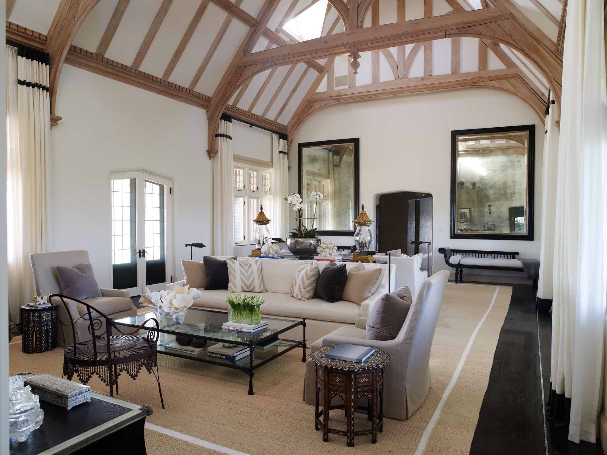 Designed by Carol Egan, this image showed the living room with vaulted ceilings and wooden oak beams.  The room has a natural abaca area rug and two seating areas with Custom Belgian sofas in "Serengeti Okapi" fabric by Donghia fabrics.  Custom Belgian slipcovered chairs in halcyon herringbone flaxen by Henry Calvin Fabrics flank a Giacometti style two-tiered coffee table with patinated bronze base and glass insert top.  There are two large antique mercury glass mirrors anchored by a pair of Anglo Raj chaise lounges flanking the opening to the Grand living room.