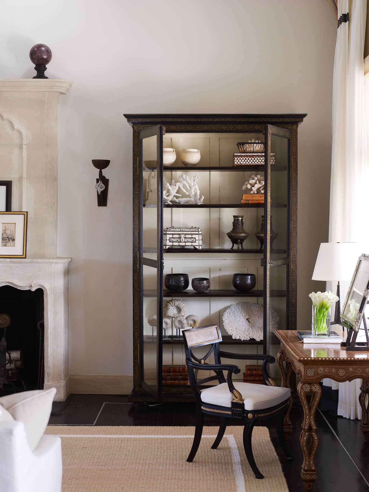 An antique displaying case flanks the Antique limestone Fireplace with limestone over mantle in this image of a detail of the living room designed by Carol Egan.  Adjacent to the curiosity cabinet displaying a collection of antique boxes and stoneware is an Antique Moroccan writing desk with a brass table lamp by Jacques Quinet.  On the tabletop is a framed drawing set into a tabletop easel by Pablo Picasso.