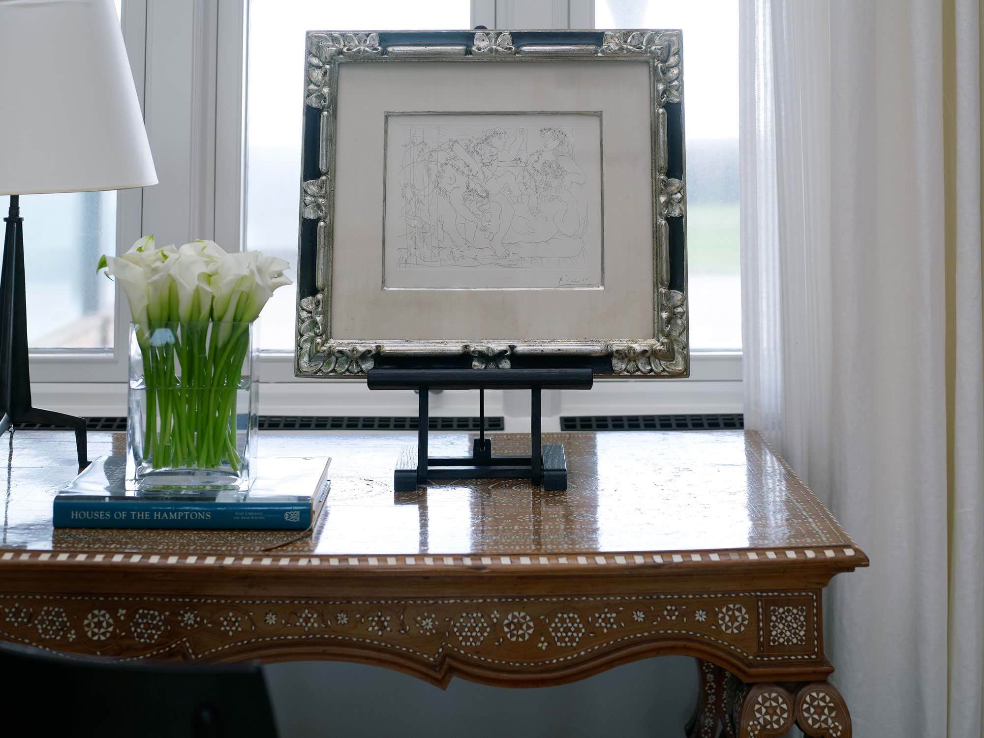 This image shows a detailed view of a Pablo Picasso drawing that is shown in a table top easel on an antique Moroccan writing table in the living room designed by Carol Egan in this Southampton Estates living room.  A brass table lamp by Jacques Quinet can be seen on the desk with a vase of French tulips.