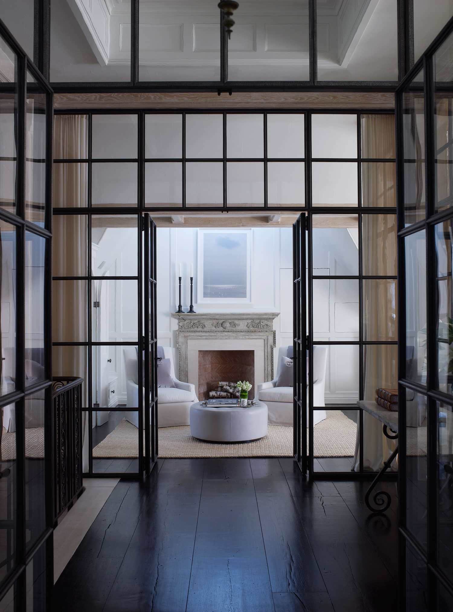 In this Image the bronze and glass vestibule frame the antique fireplace in the master bedroom suite of the Southampton residence designed by Carol Egan.  Custom Belgian chairs in Sicily texture in cream & rope by Henry Calvin fabrics flank the fireplace with an ottoman detailed with biscuit tufting.