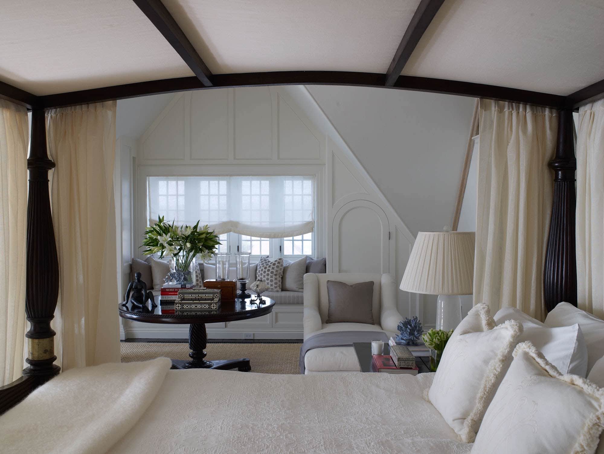 An antique Anglo Raj tester bed is upholstered in white linen in the master bedroom designed by Carol Egan and shown in this image.  The Bed canopy and canopy curtains in "Meghan" sheer ivory fabric by Lee Jofa frame the image.  "Coolie" shaped, pleated lampshades in "Gossamer Gauze" fabric by Rogers & Goffigon are seen on the crystal table lamps on the bedside tables.  Two Belgian style Chaise Lounges flank an antique Center wooden table in the window which overlooks the ocean.