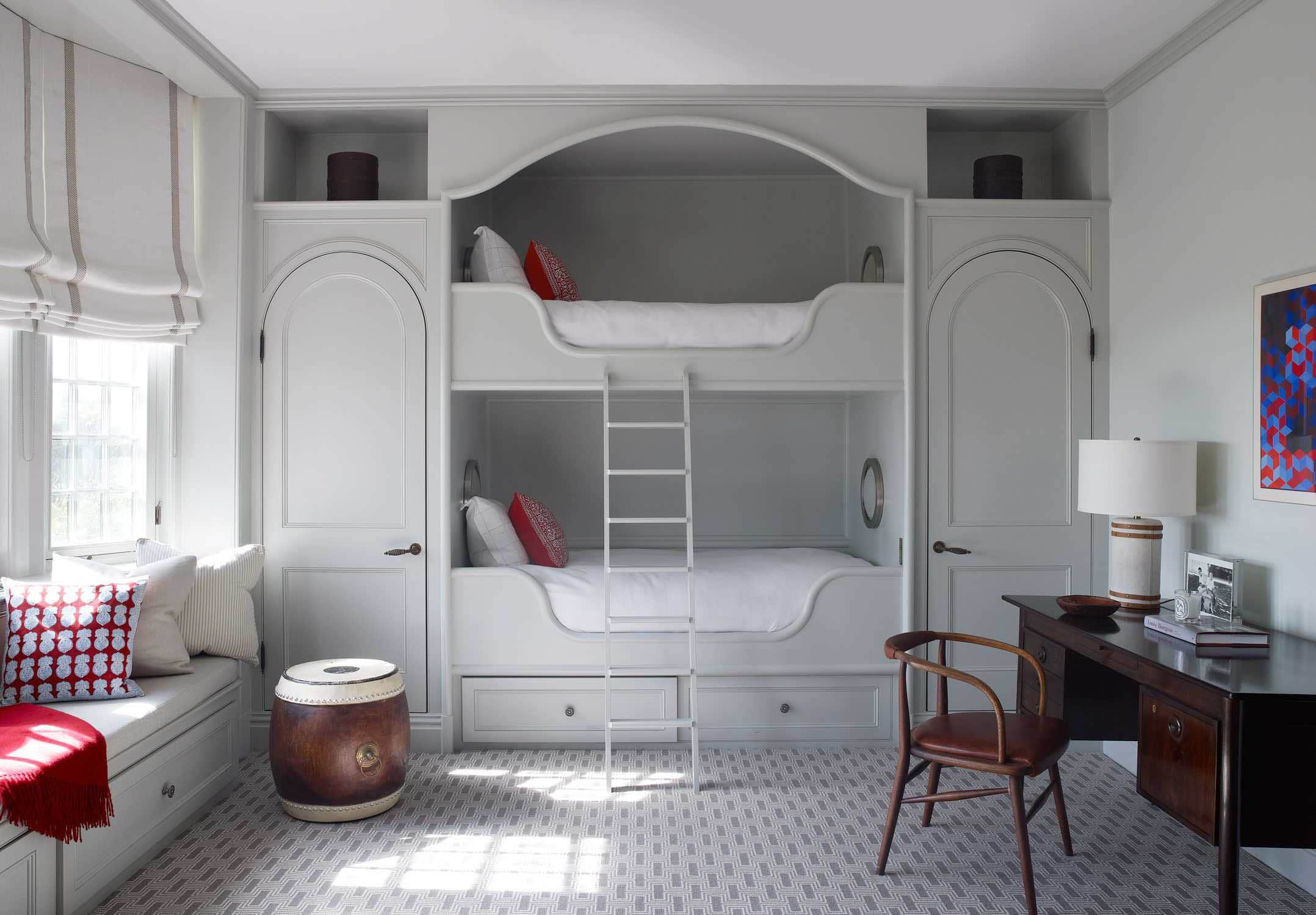 Designed by Carol Egan, this interior image shows a guest bedroom for children with painted grey bunk beds.  The room has a wall to wall carpet in natural wool and an vintage danish writing table is adjacent to the bunk beds.  There is a window seat upholstered and accented with batik printed throw pillows.