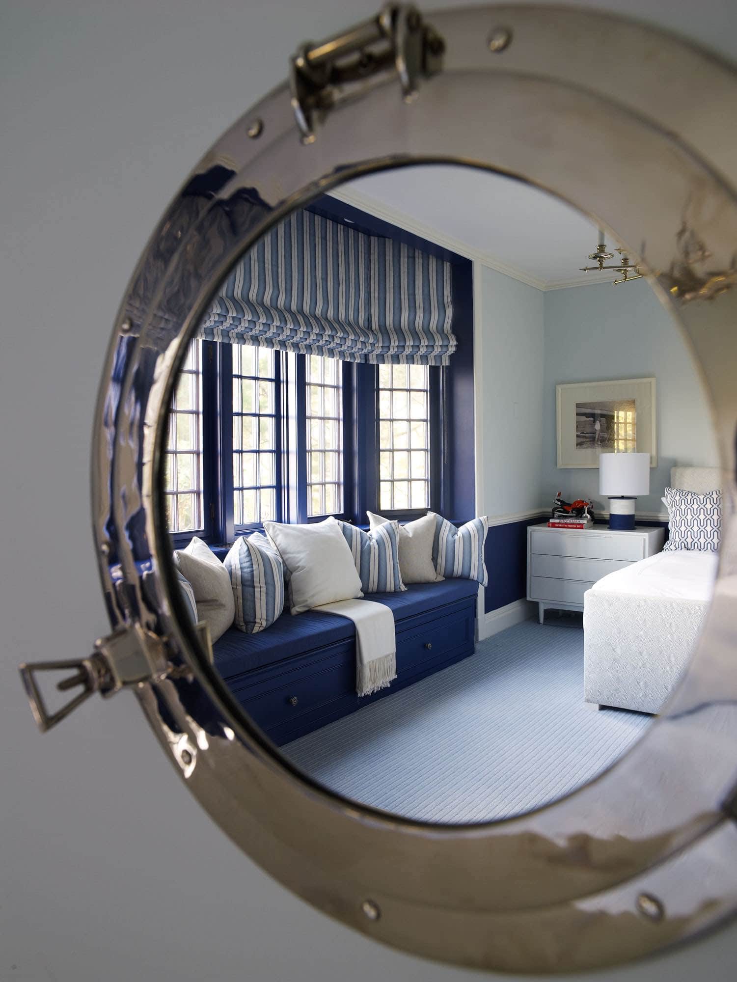 Shown in the reflection of a polished nickel portal mirror is a boy’s bedroom designed by Carol Egan in this Southampton residence.  The millwork in the room is painted in a Wainscott navy blue and accented with pale blue walls. A trundle bed by Charles H. Beckley is accessorized with throw pillows in Gorrivan Fretwork in navy periwinkle by China Seas fabric House.  The window seat is upholstered in a Rogers & Goffigon linen and the roman shade fabric is a Stripe in ocean blue.